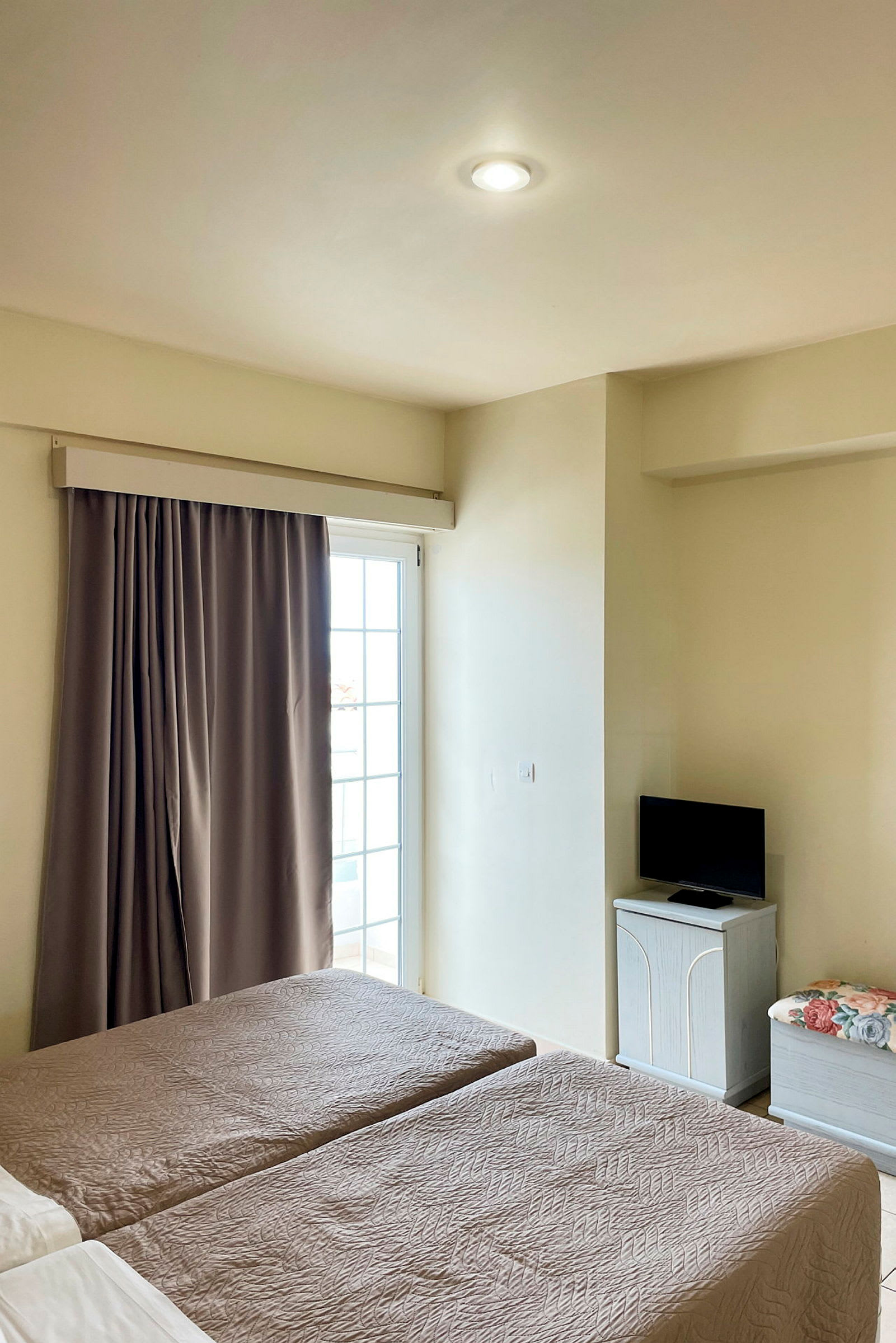 Ett hotellrum med två enkelsängar, en balkongdörr med gardin, och en liten TV på ett skåp. (AI-genererad text)