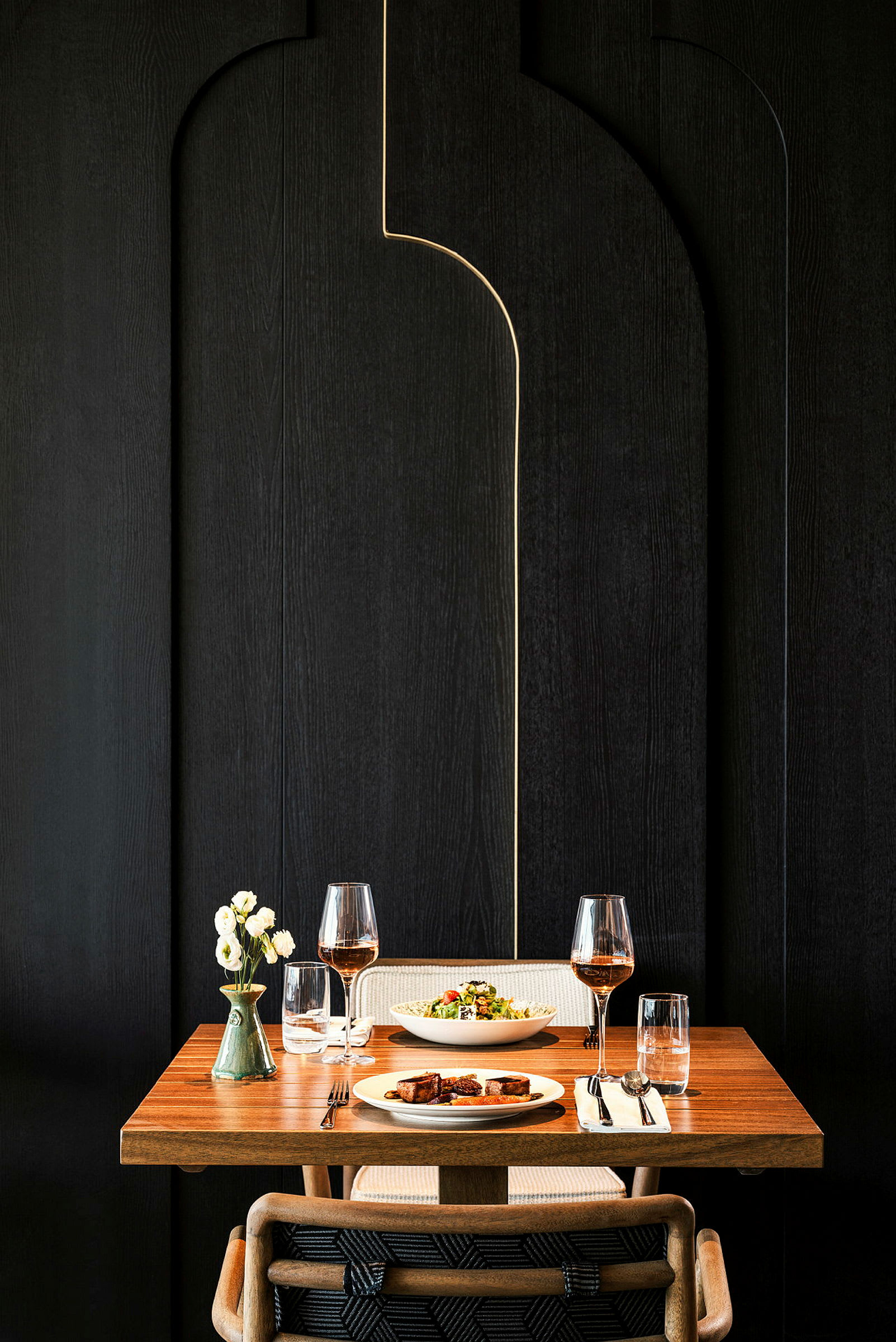 Ett dukat bord med en träyta, för två personer, två glas vin, två tallrikar med mat och en vas med blommor. (AI-genererad text)
