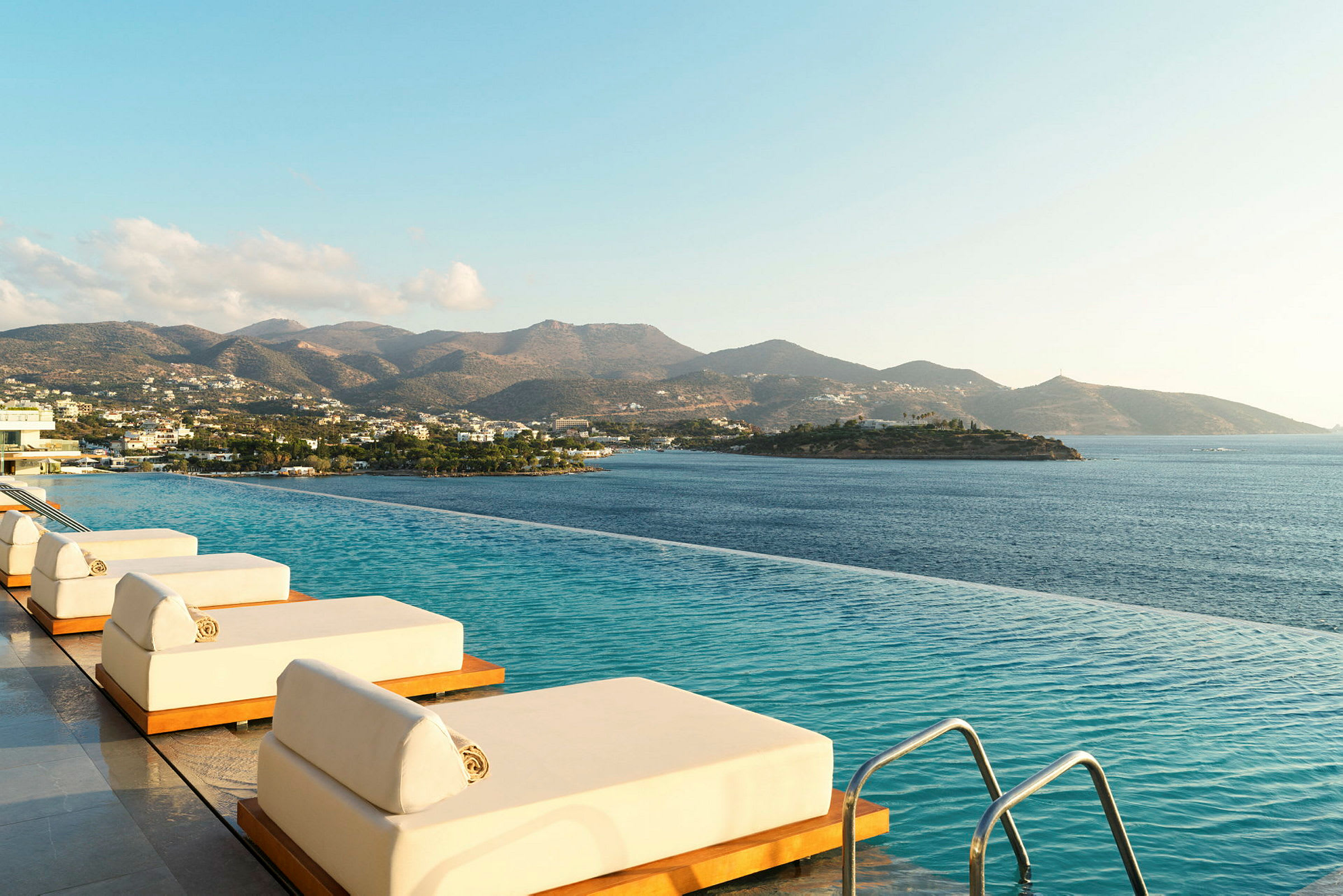 Inifinitypool med solstolar vid havet och berg i bakgrunden. (AI-genererad text)
