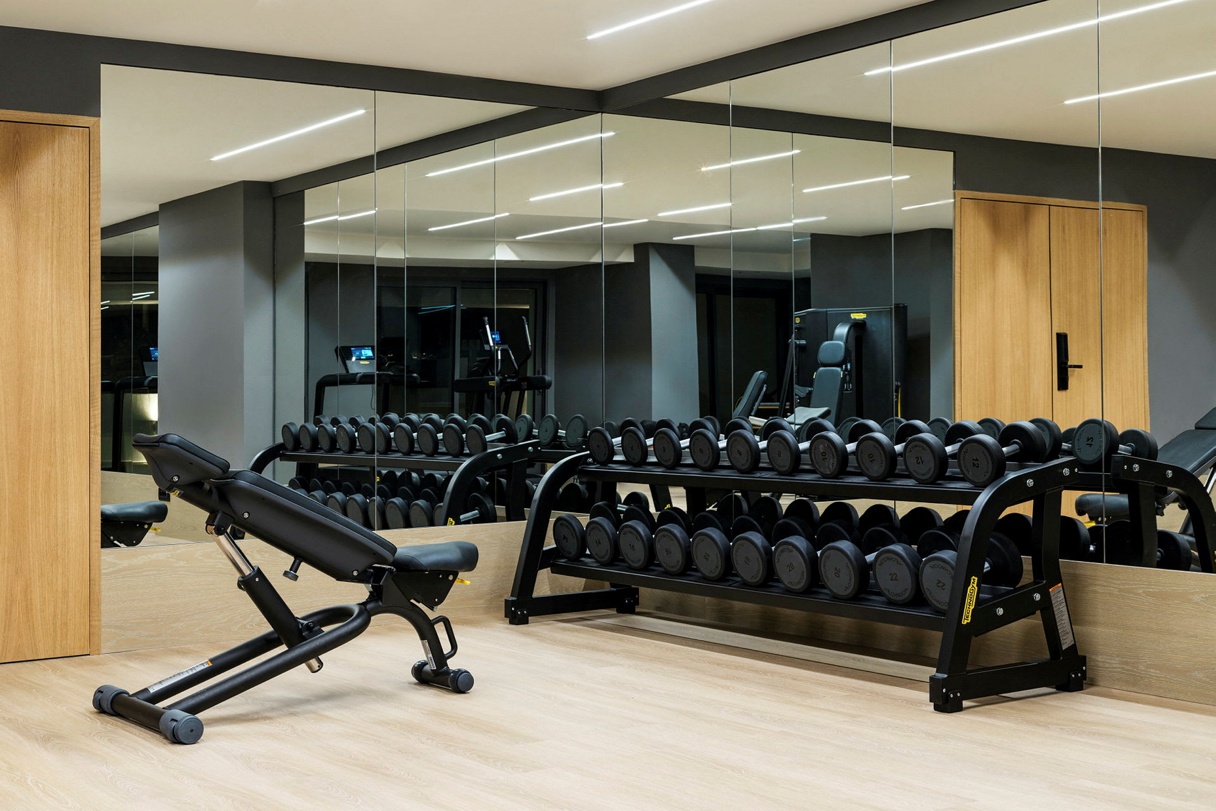 Ett gym med hantelställ och träningsbänk, omgiven av speglar och grå väggar. (AI-genererad text)
