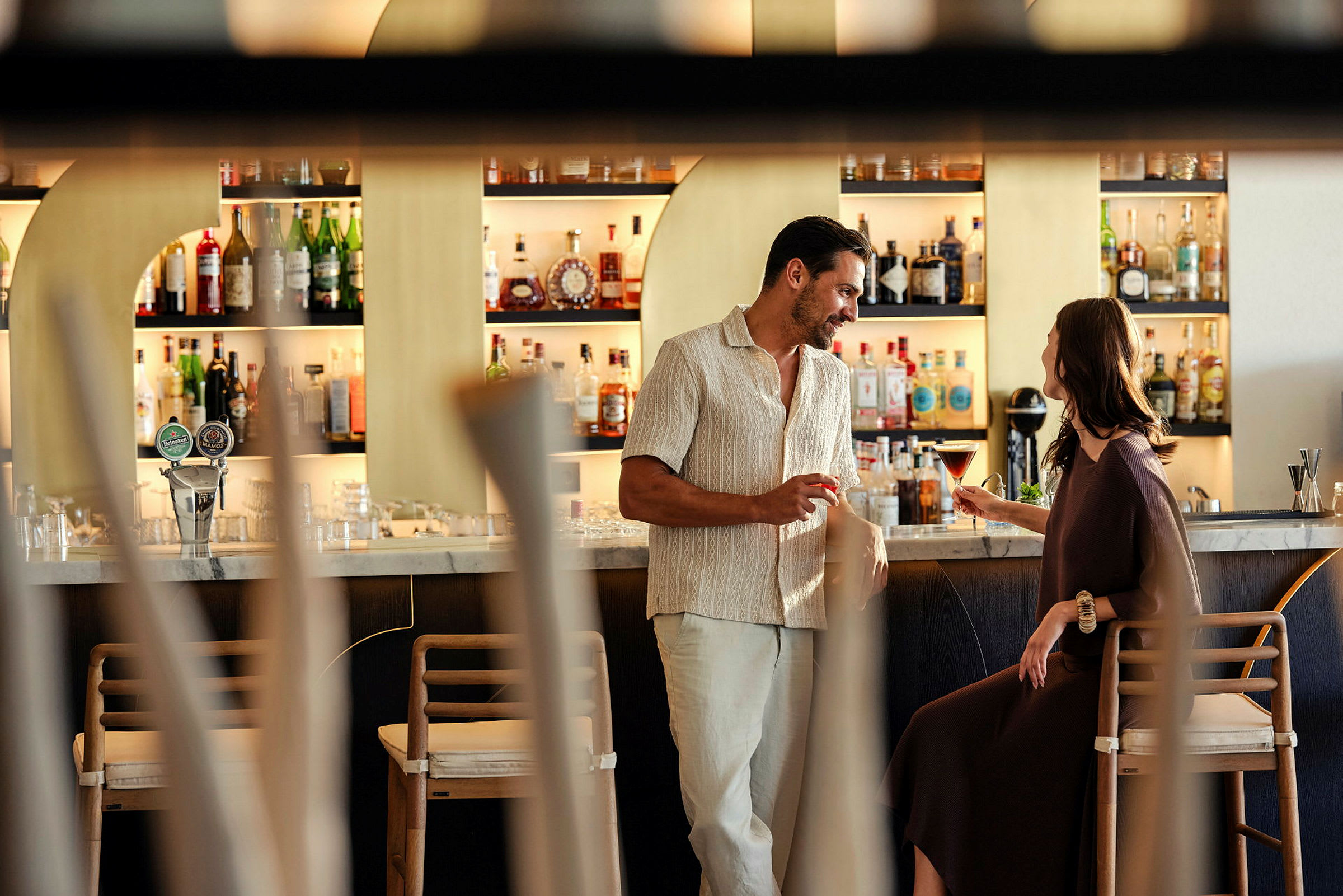 Två personer samtalar vid en modern bar med olika drycker i bakgrunden. (AI-genererad text)