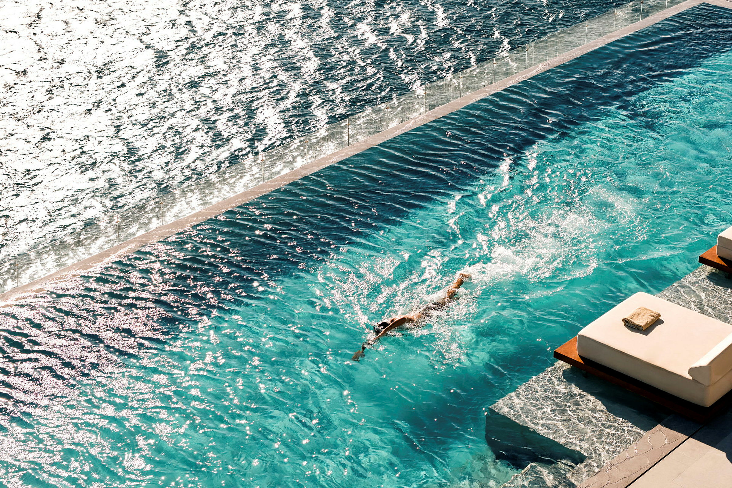 En person simmar i en pool vid sidan av havet, med solstolar i förgrunden. (AI-genererad text)