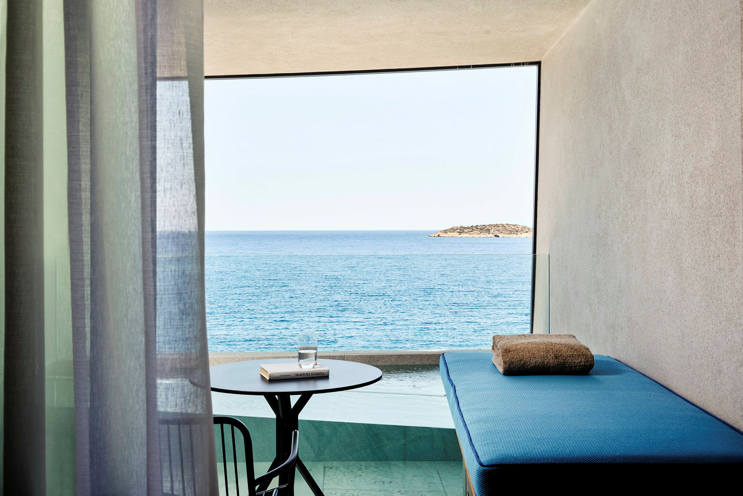 En balkong med utsikt över havet och en ö i fjärran. På balkongen står ett litet bord med ett glas vatten och en bok samt en blå solbädd med en handduk. (AI-genererad text)