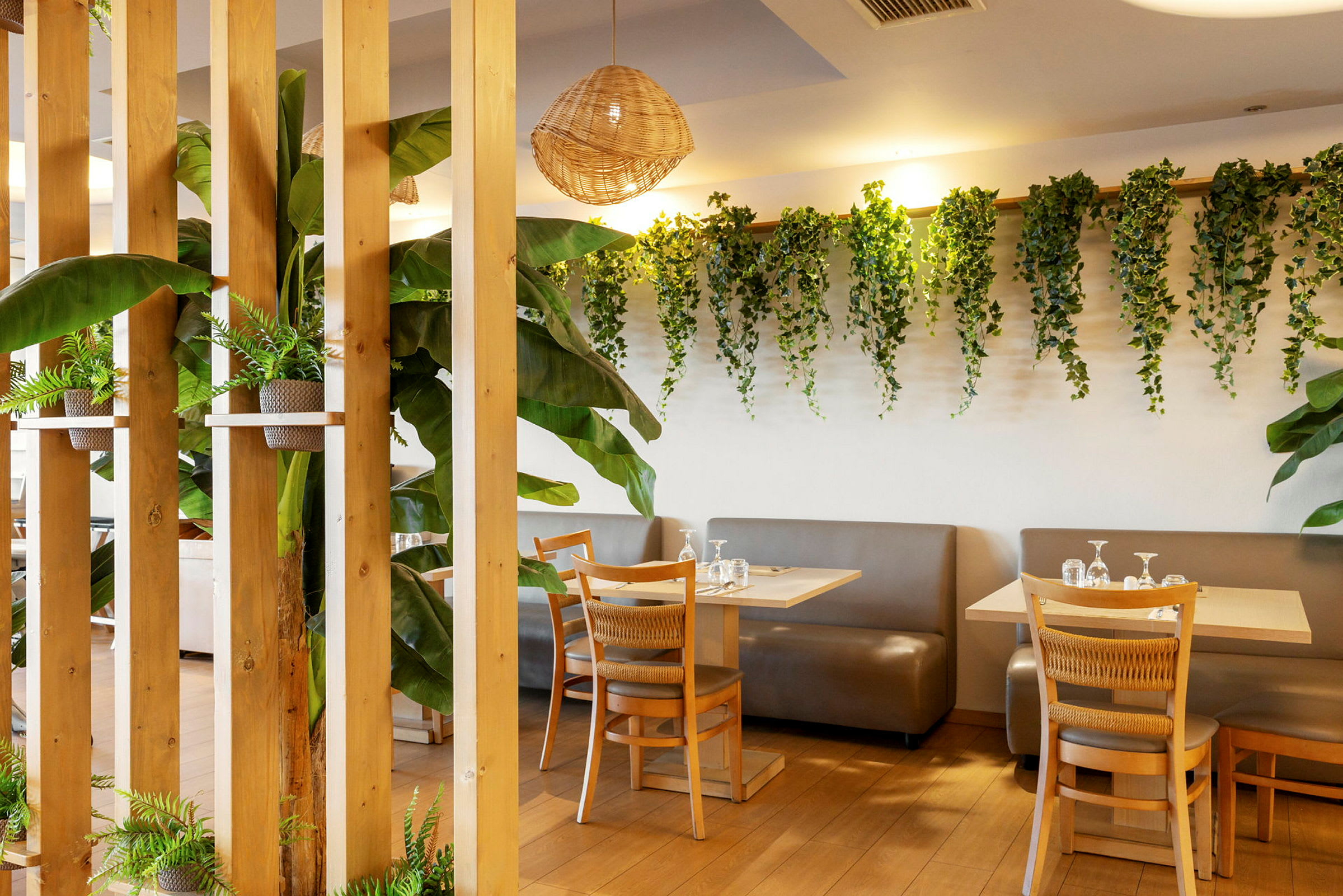 En inomhusrestaurang med bord och stolar i trä. Väggarna är dekorerade med gröna växter, och det finns betydande inslag av gröna växer runt omkring. (AI-genererad text)