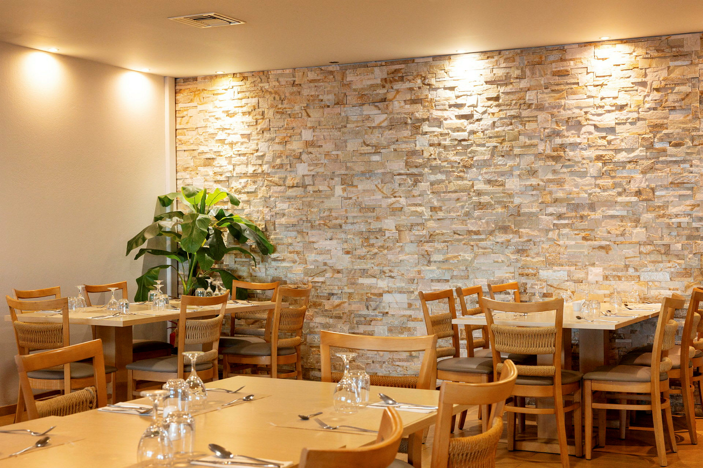Restaurangmiljö med bord och stolar framför en vägg av sten. En grön växt står i hörnet. (AI-genererad text)