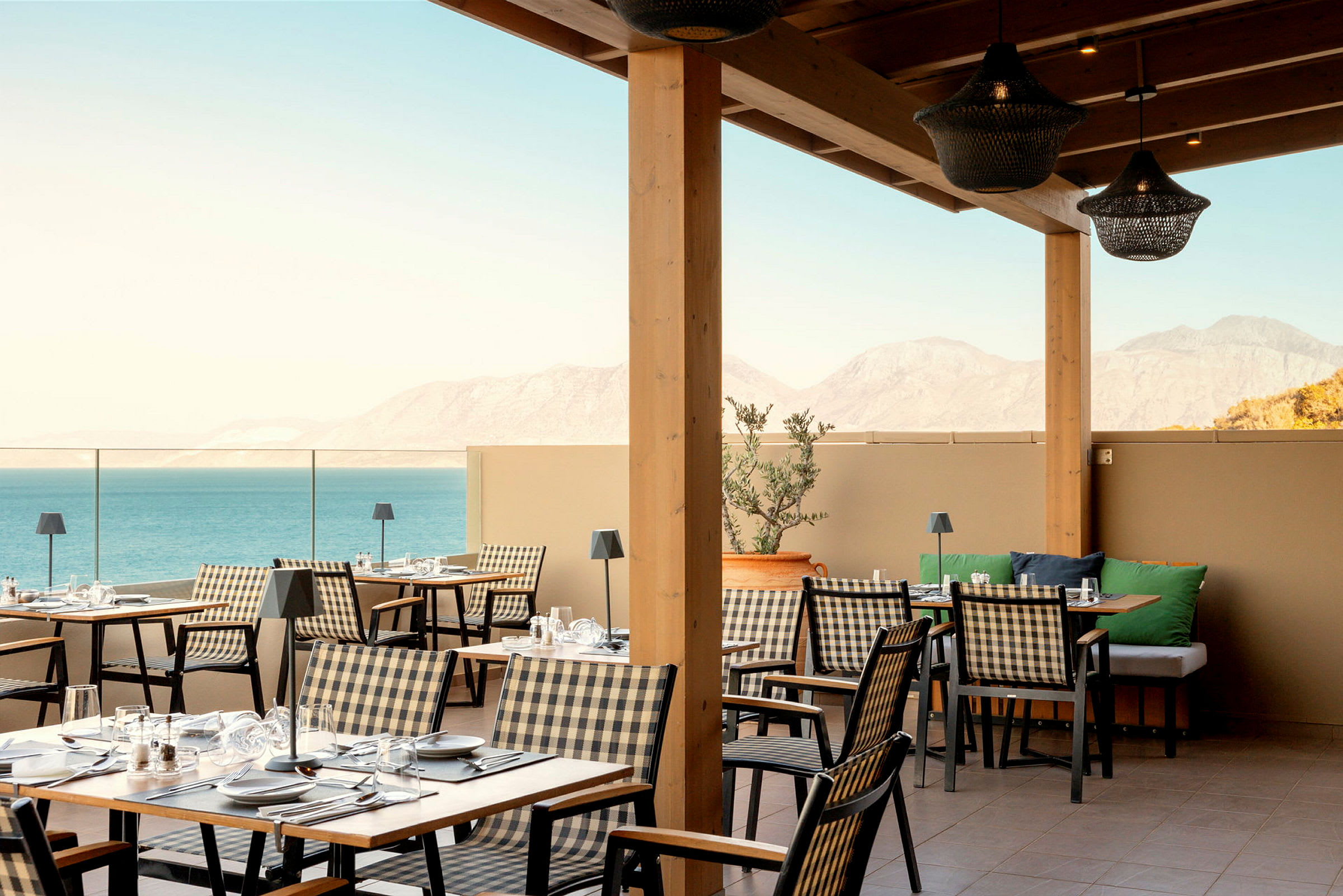 En utomhusrestaurang med rutiga stolar och träbord, och en utsikt över havet och bergen. (AI-genererad text)