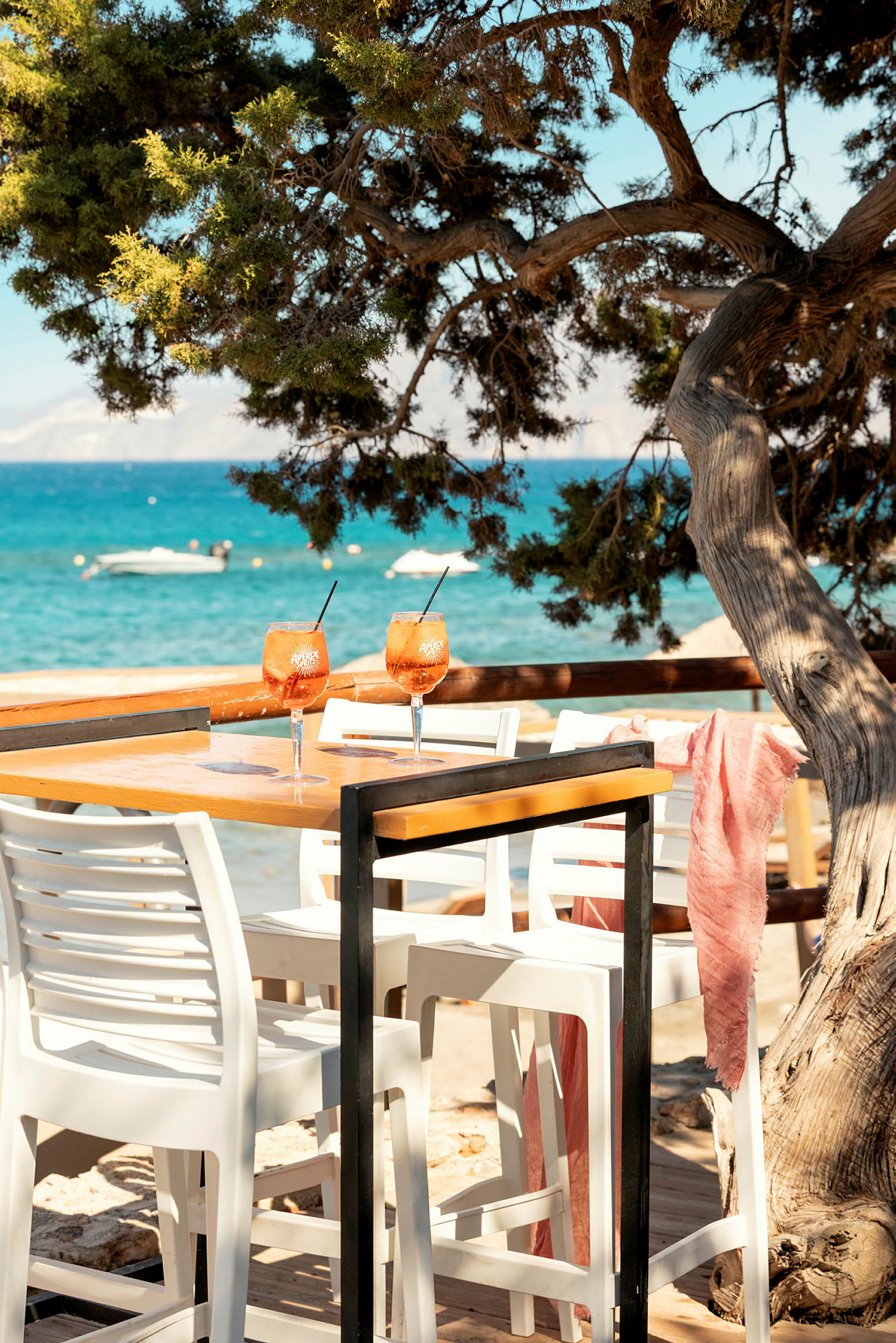 Ett träbord med vita plaststolar framför ett träd, med utsikt över havet och båtar i bakgrunden. På bordet står två glas med orange dryck. (AI-genererad text)