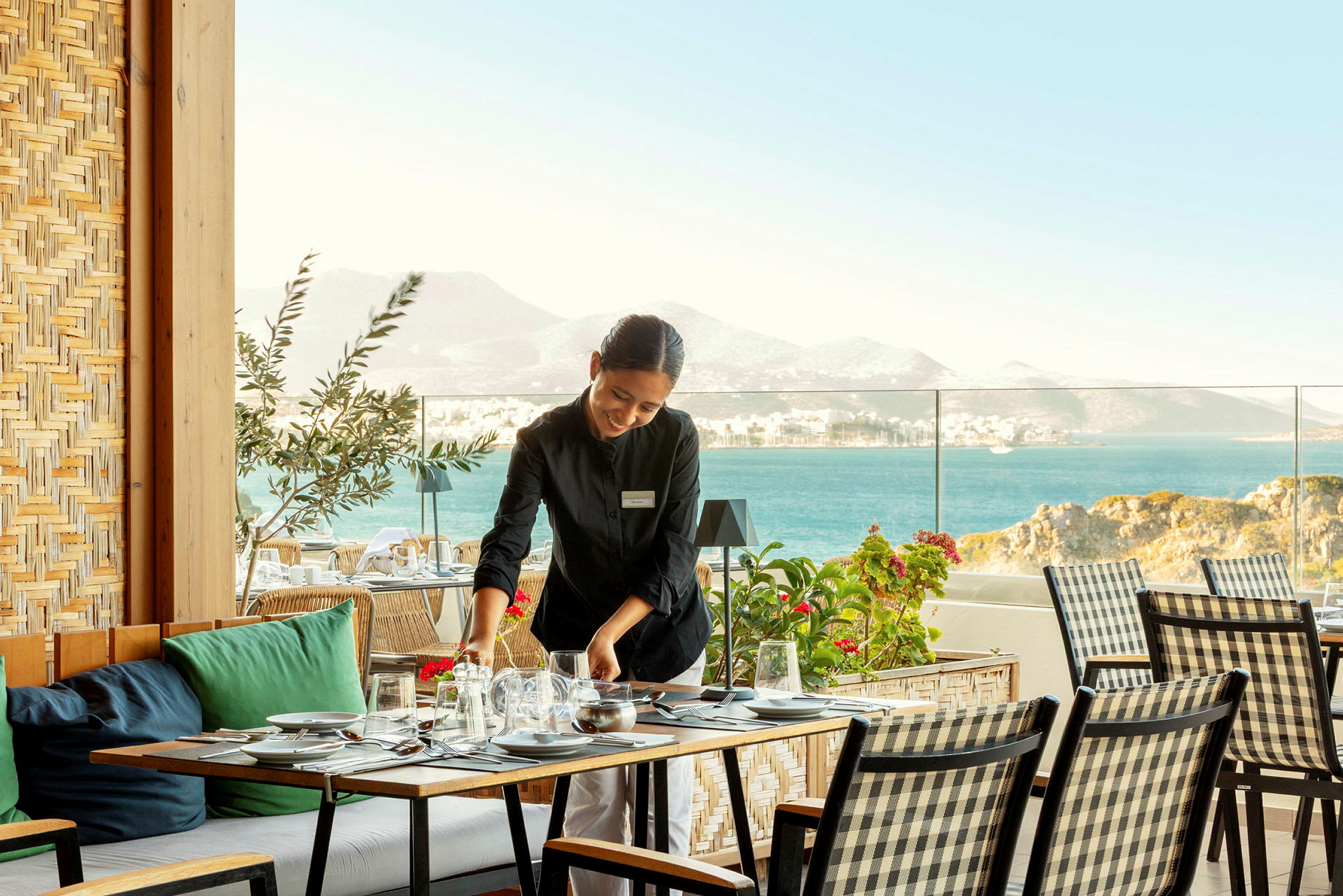 En servitör dukar ett bord på en terrass med havsutsikt och berg i bakgrunden. (AI-genererad text)
