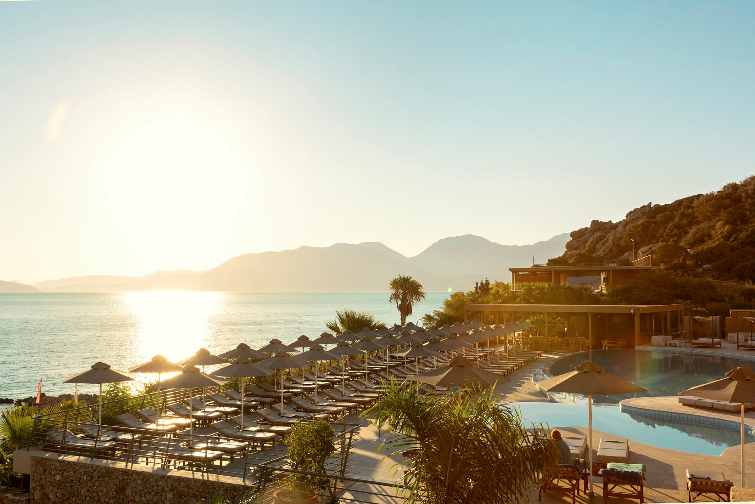 Solnedgång vid havet där solstolar och parasoller står vid en pool och en terrass. (AI-genererad text)