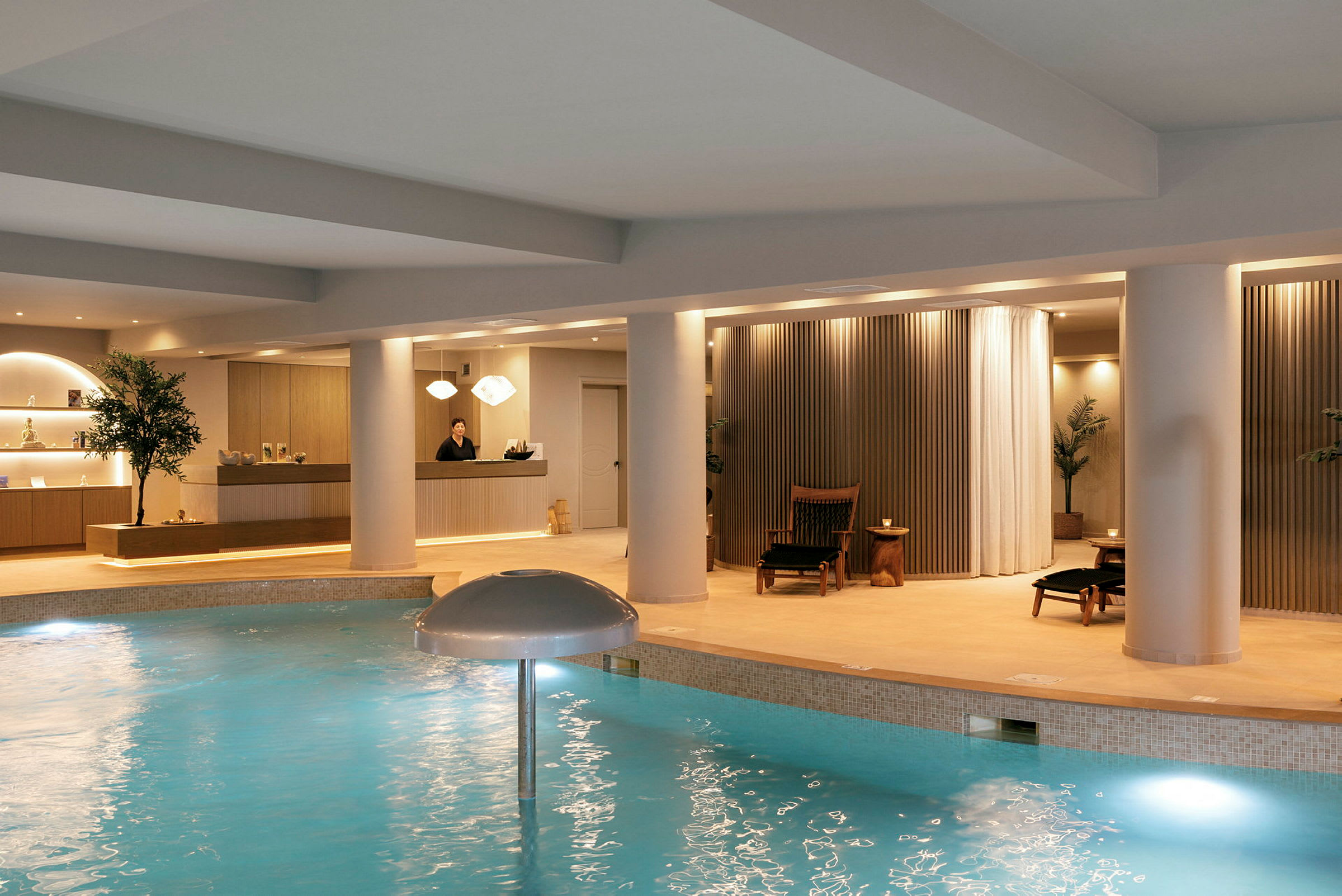 Ett modernt spa med en inomhuspool, reception och avkopplingsområden. En person står i receptionen. (AI-genererad text)