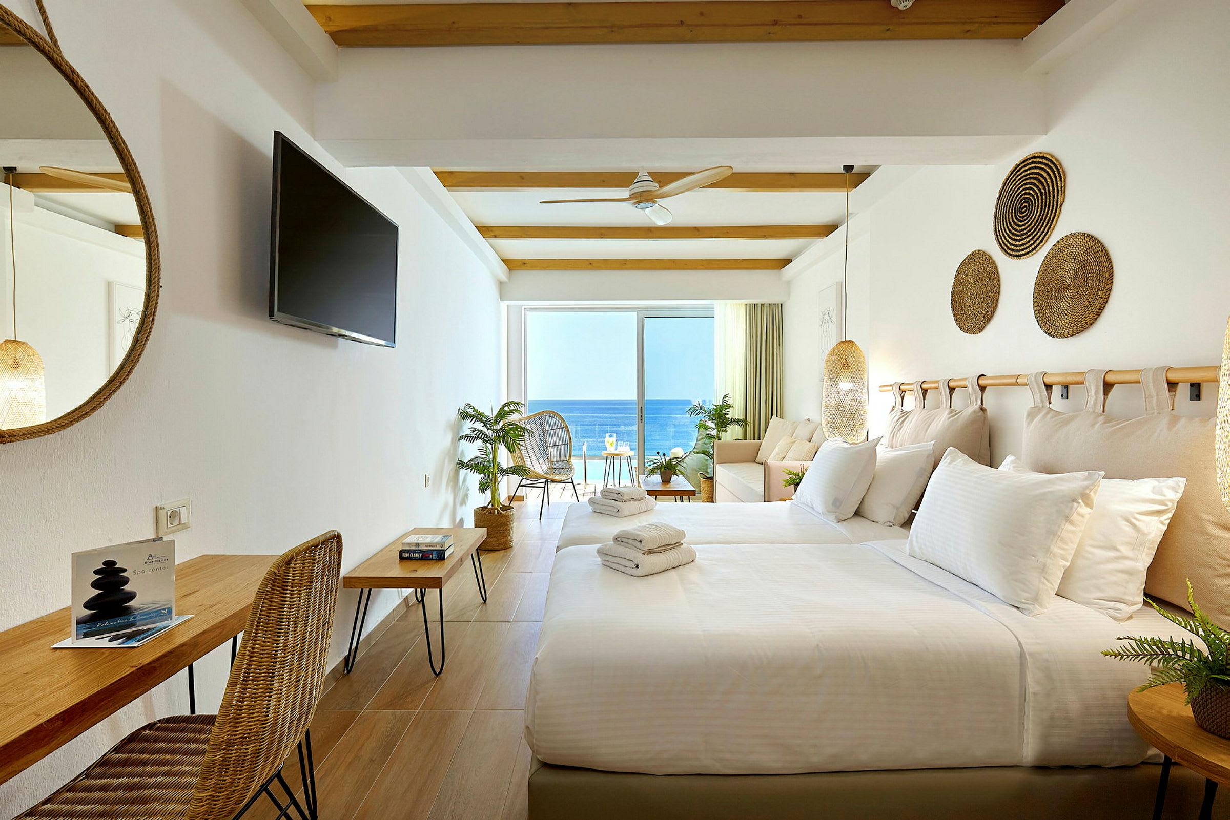Ett hotellrum med vita väggar, en bäddad säng, sittmöbler och en balkong med utsikt över havet. (AI-genererad text)