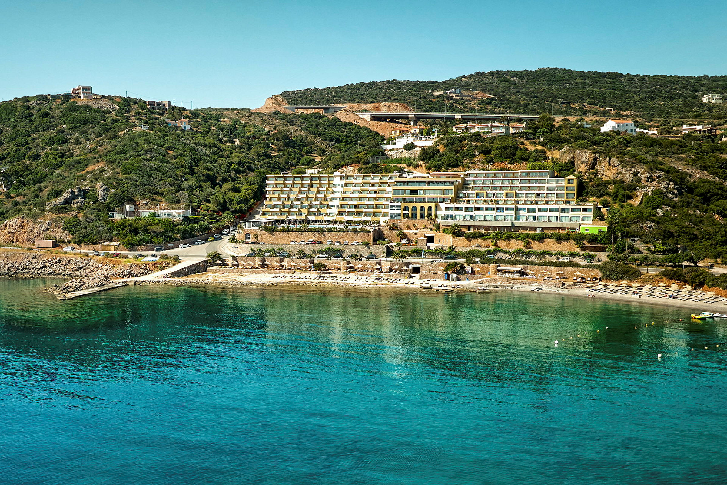Hotell vid strand med klart blått vatten och grönskande kullar i bakgrunden. (AI-genererad text)