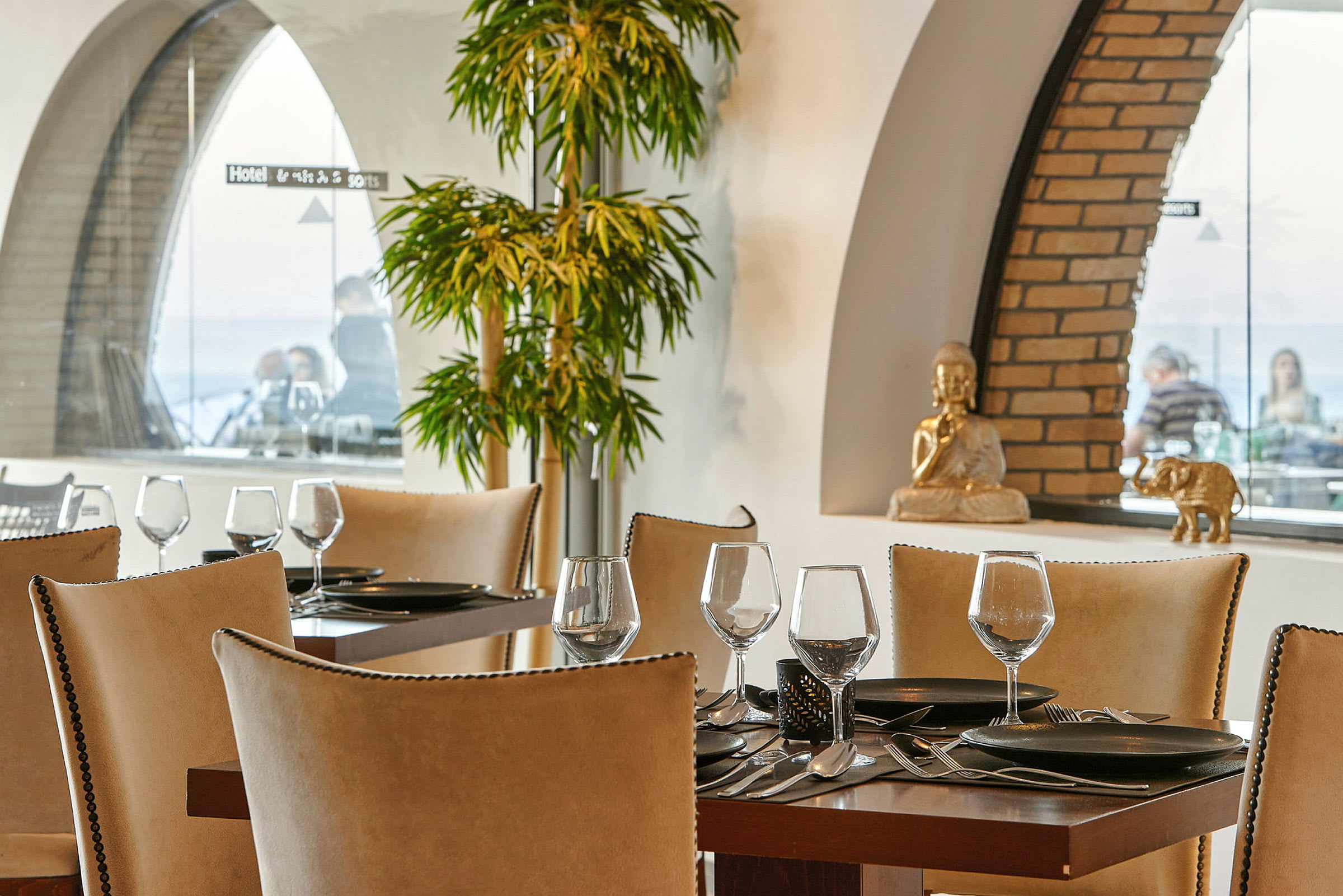 Ett restaurangbord med uppdukad servis och glas, omgiven av elegant inredning med gröna växter och dekorativa figurer i bakgrunden. (AI-genererad text)