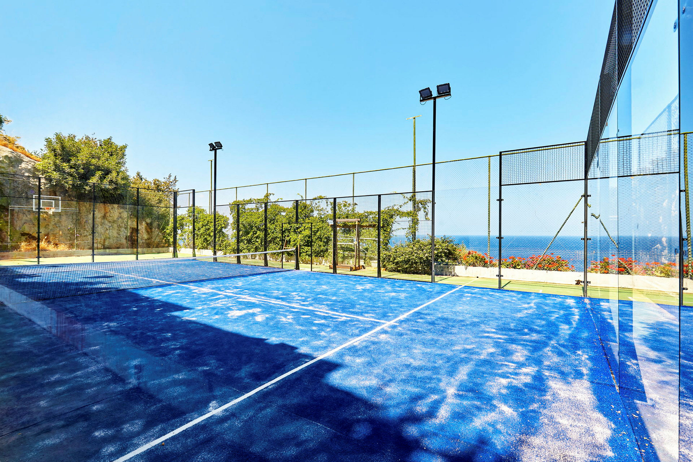 En padelbana med blått underlag omgiven av stängsel med havsutsikt. (AI-genererad text)