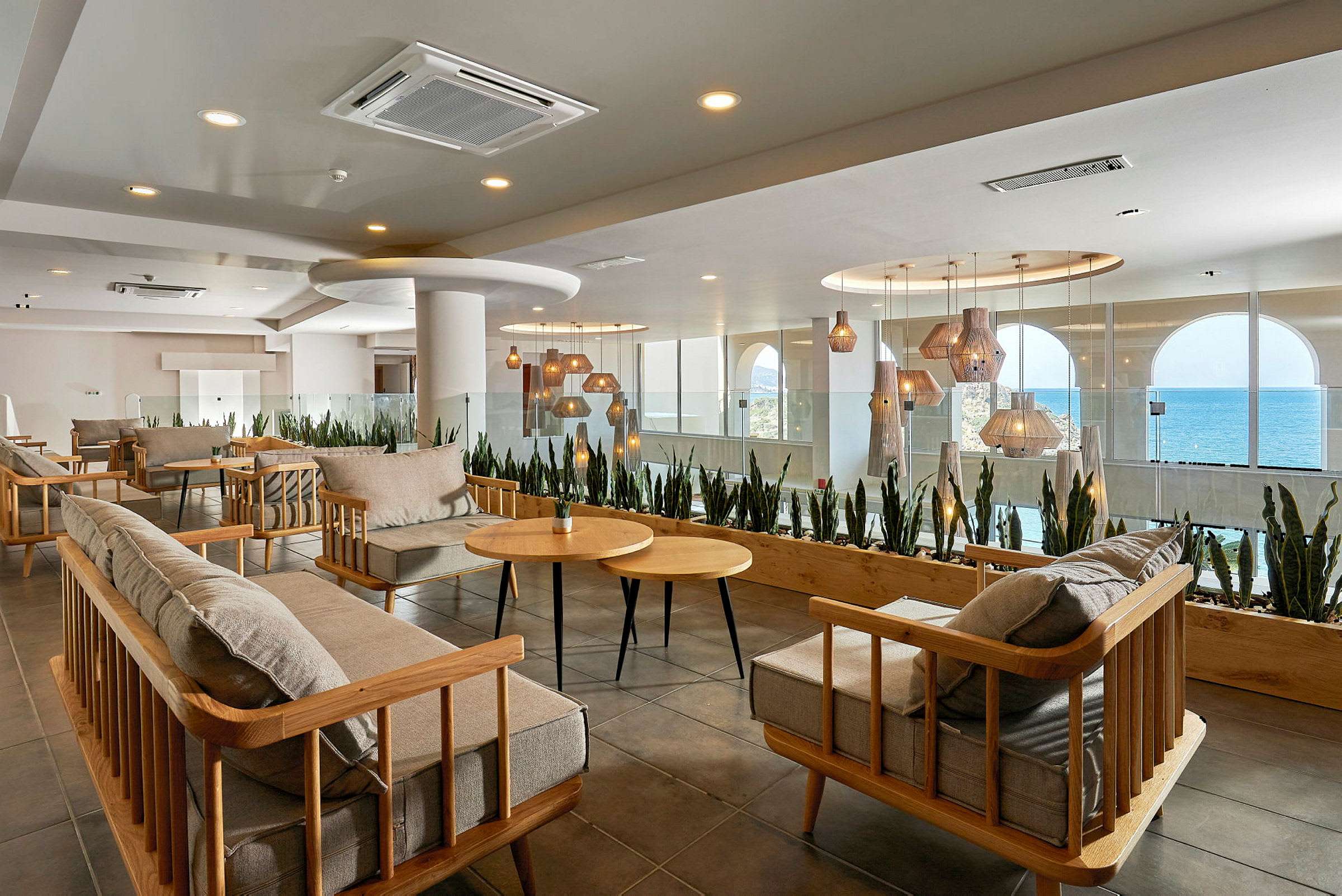 En modern lounge med trästolar och bord samt hängande lampor. Stora fönster med utsikt över havet i bakgrunden. (AI-genererad text)