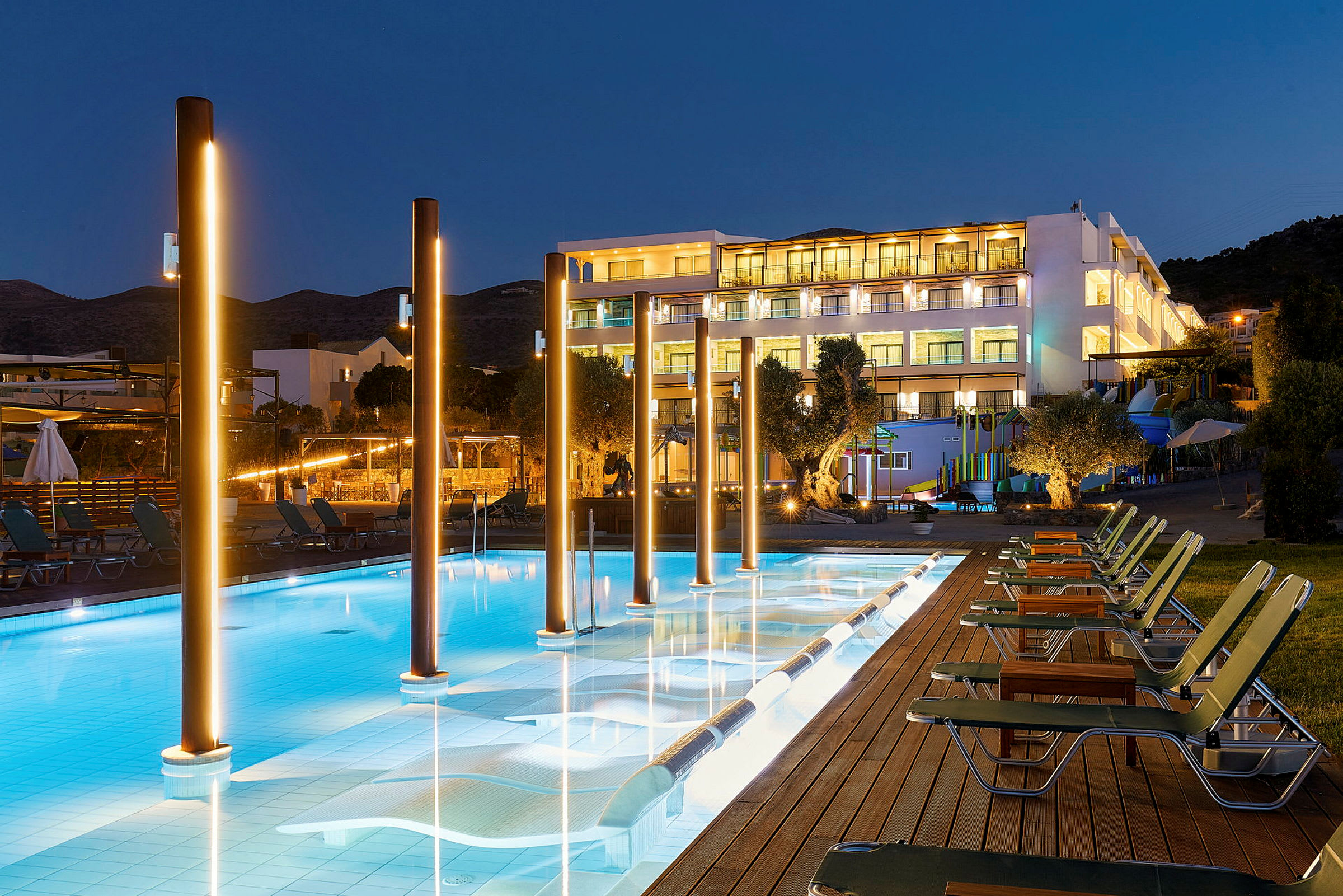 En upplyst hotellfasad bakom en pool med stolar vid sidan, fotograferad på kvällen. (AI-genererad text)