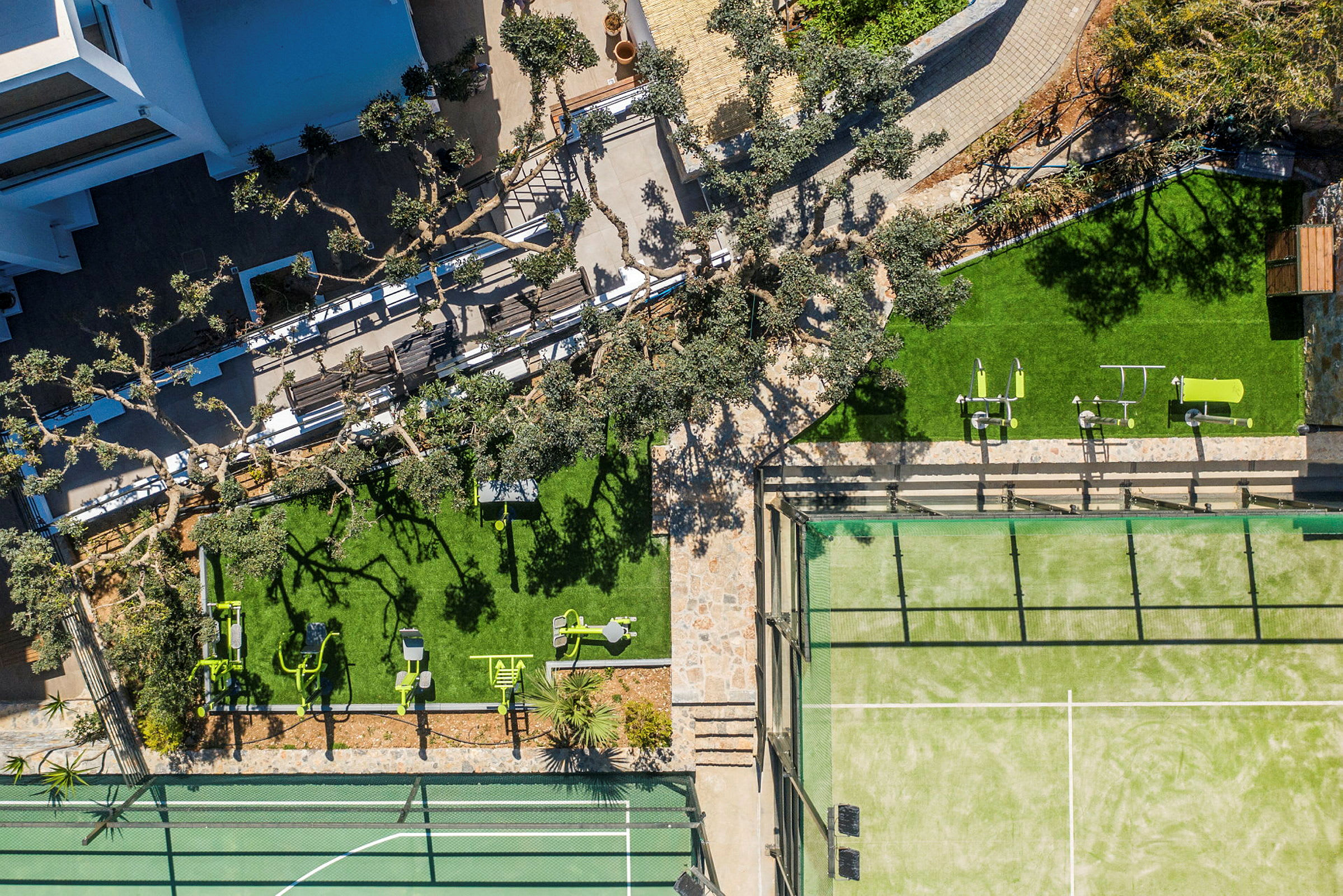 Flygfoto av en utomhusmiljö med träd, gröna gräsmattor, fitnessutrustning och en tennisbana. (AI-genererad text)