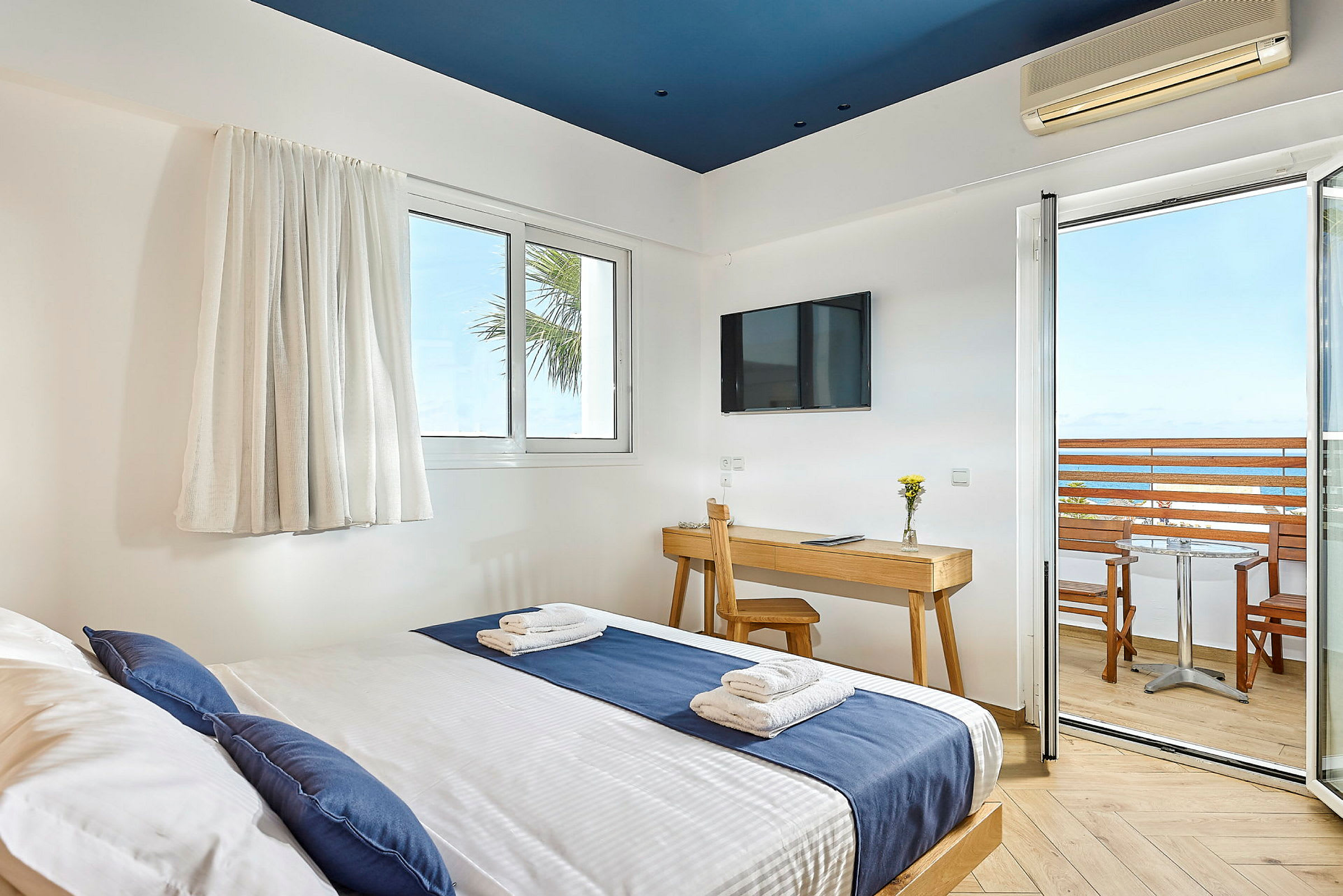 Hotellrum med säng klädd i blå och vit bäddning, skrivbord med stol, TV på väggen, och balkong med utsikt över havet. (AI-genererad text)