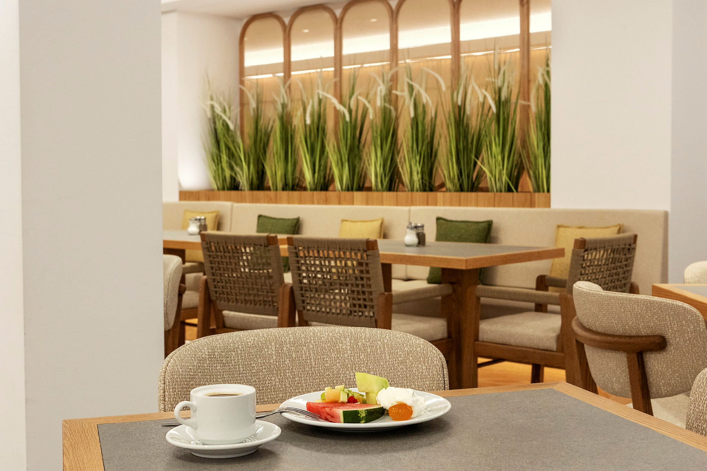 Bild av en restaurang med bord, stolar och gröna växter som dekor. I förgrunden syns en kaffekopp och en tallrik med frukt. (AI-genererad text)