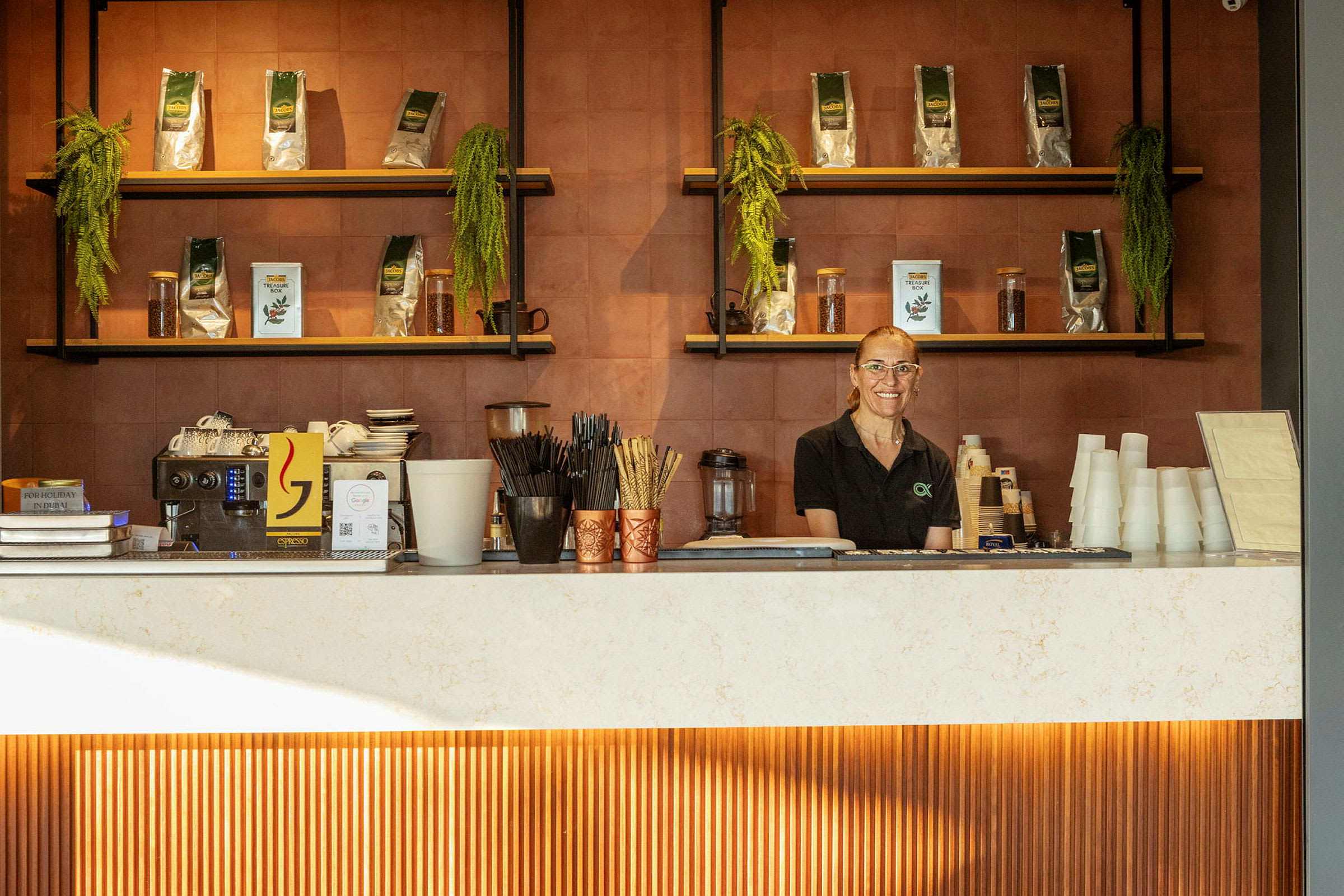En person står bakom en disk i ett kafé, omgiven av hyllor med kaffeprodukter och dekorationer. (AI-genererad text)