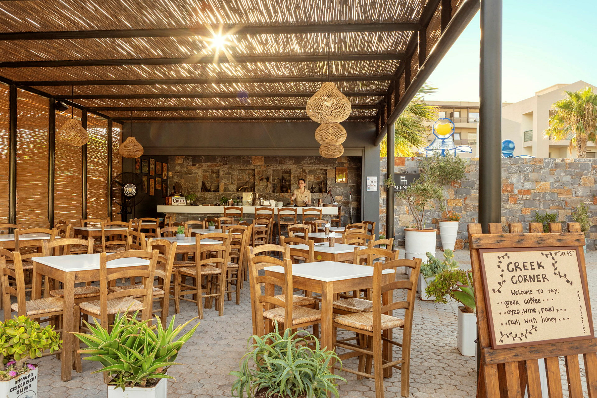 Här syns en utomhusrestaurang med träbord och stolar under ett tak av bambu, samt en skylt med texten "Greek Corner". (AI-genererad text)