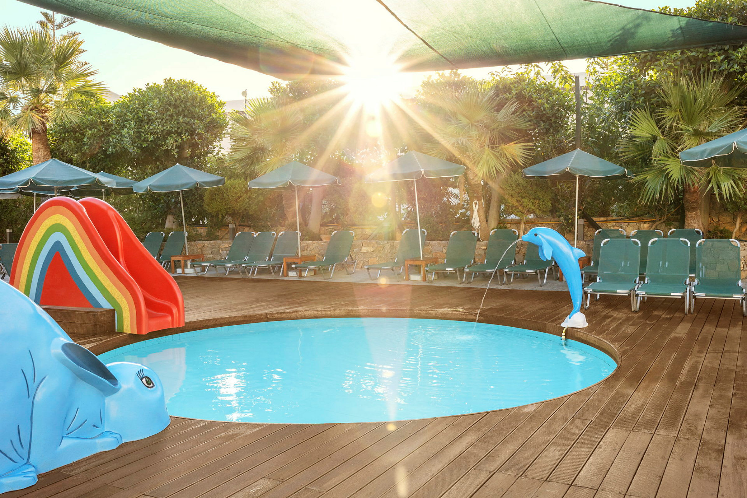En liten rund barnpool omgiven av ett trädäck. I poolen finns en blå delfinfigur som sprutar vatten och en rutschkana i regnbågens färger. Solstolar och parasoller står runt poolen. (AI-genererad text)
