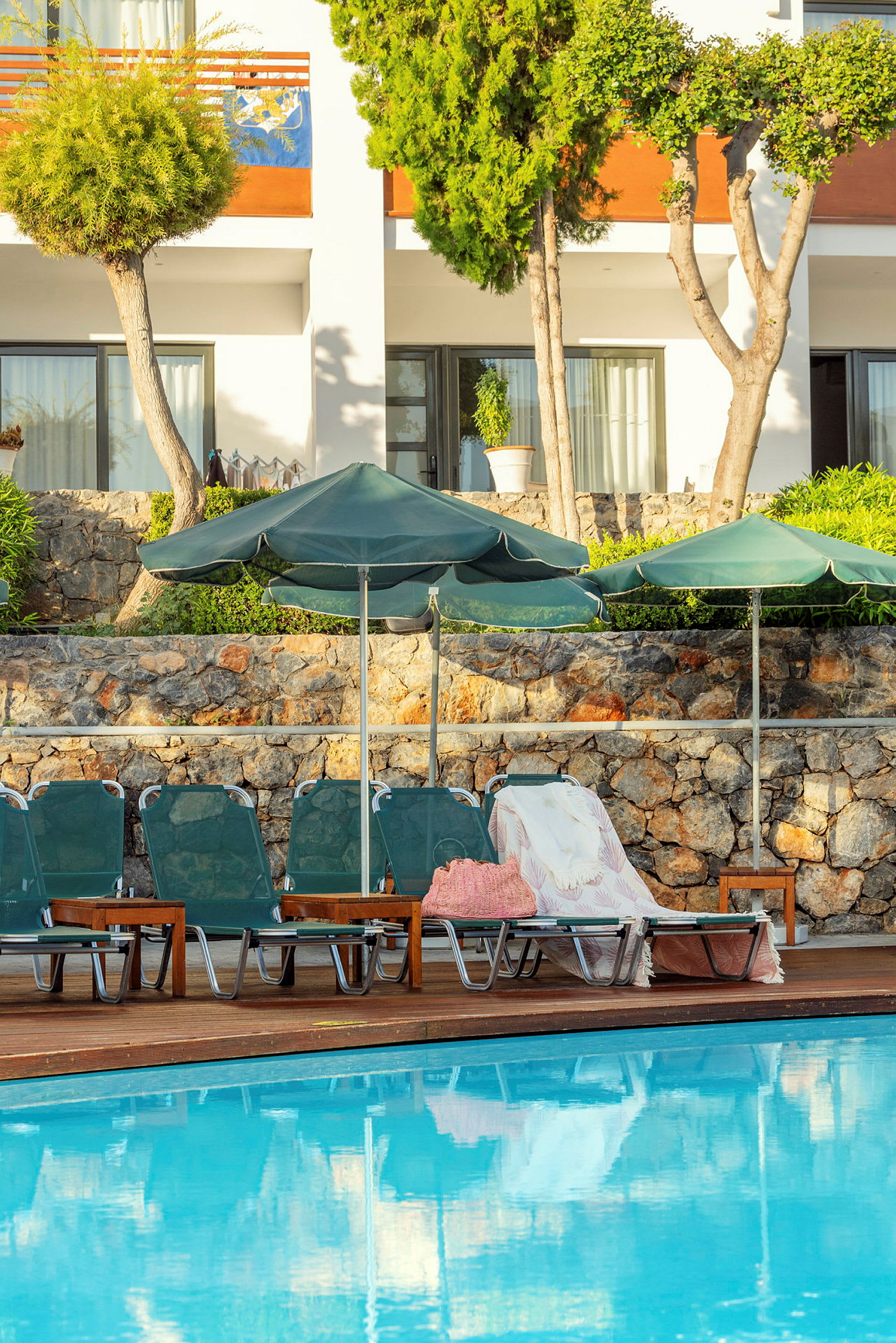 Bild av en pool med solstolar och parasoller framför en byggnad omgiven av grönska. (AI-genererad text)