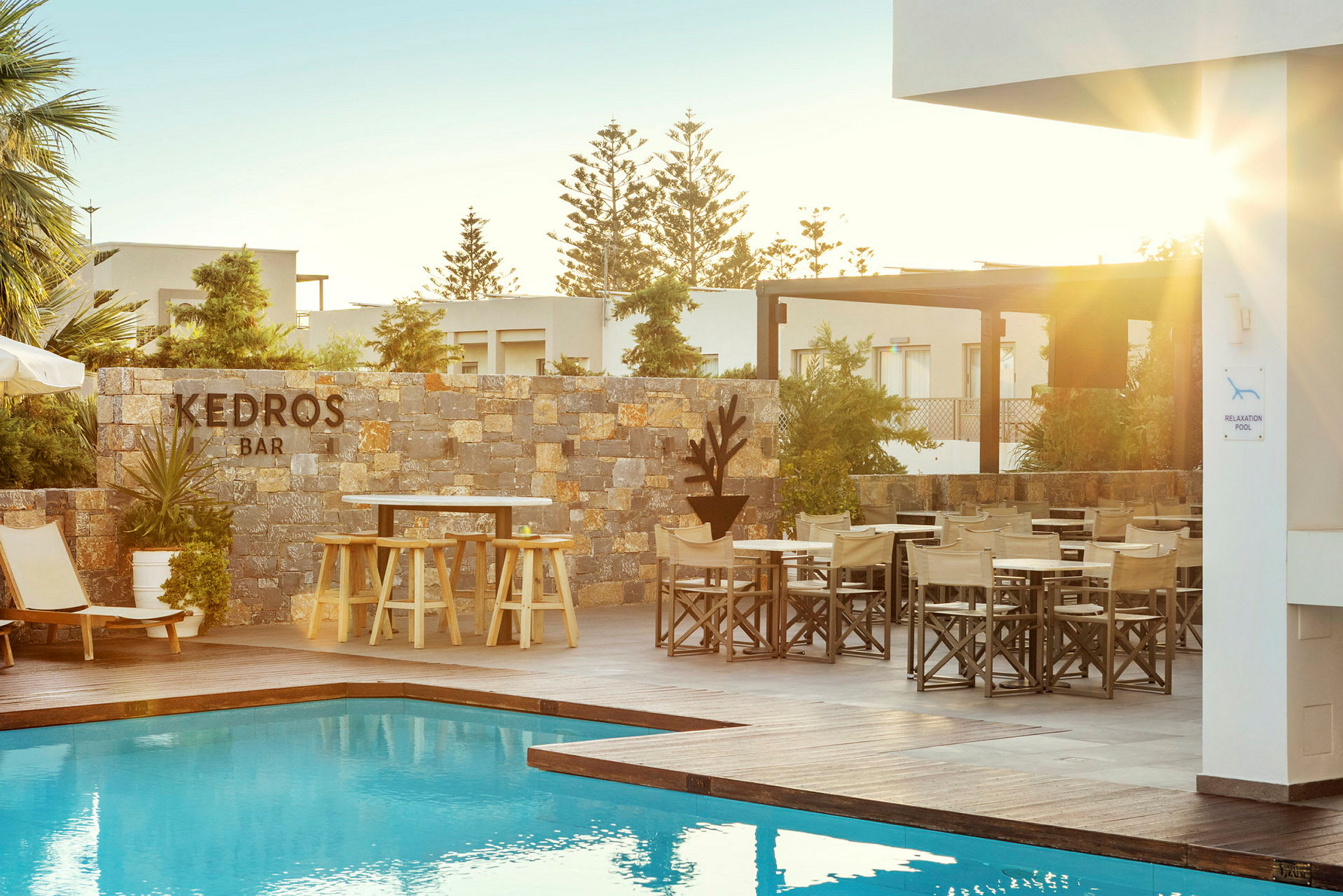 En utomhuspool med solstolar och bord bredvid en stenmur märkt "Kedros Bar". Solen skiner över området. (AI-genererad text)
