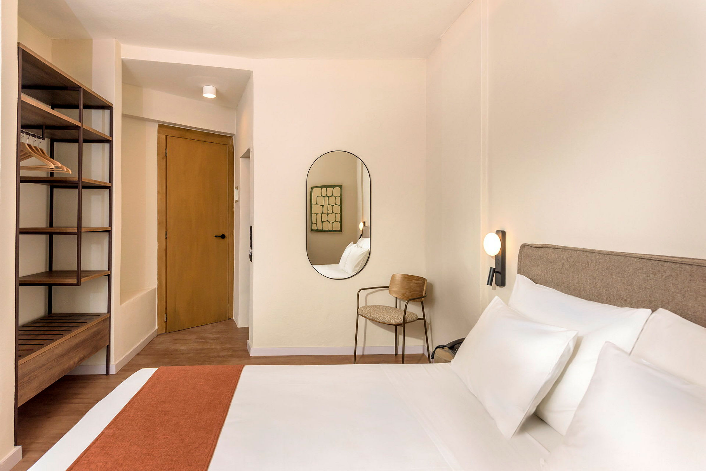Ett hotellrum med en säng, en spegel, en stol och en öppen klädförvaring. (AI-genererad text)