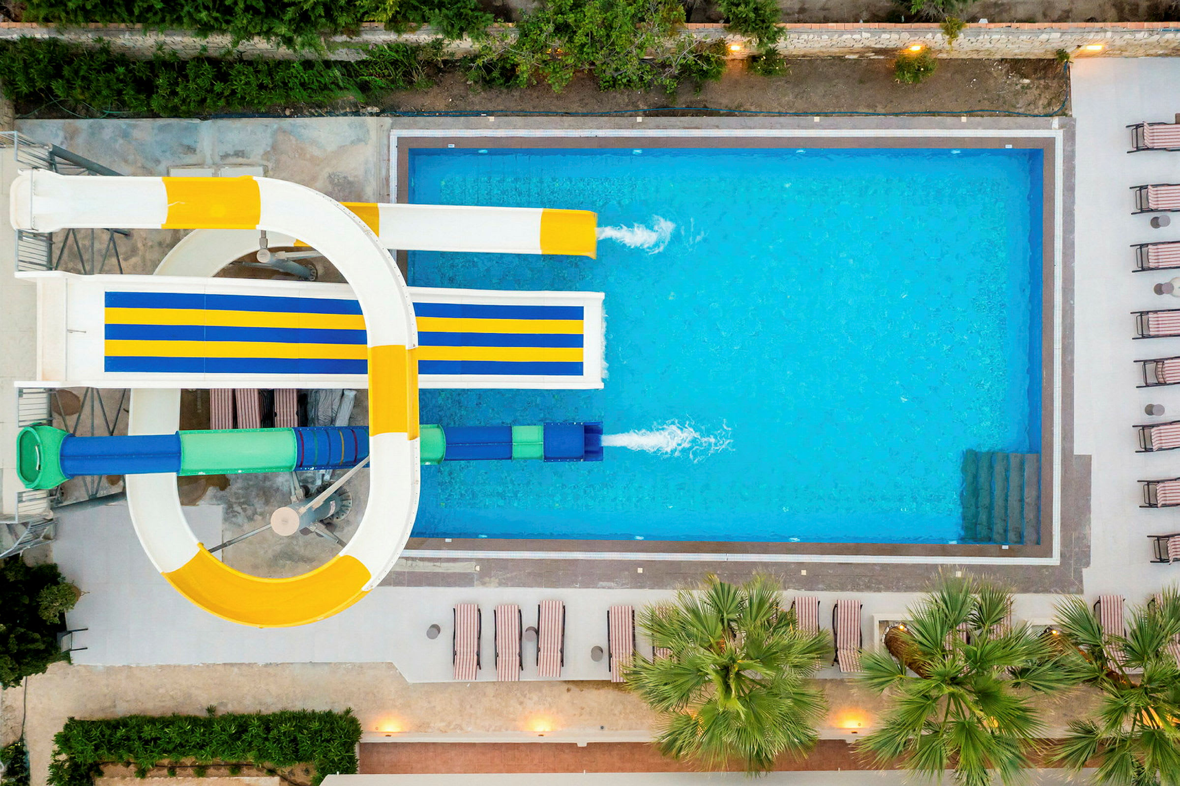 Färgglada vattenrutschbanor som leder ner i en klarblå pool, omgiven av solstolar och palmer. (AI-genererad text)