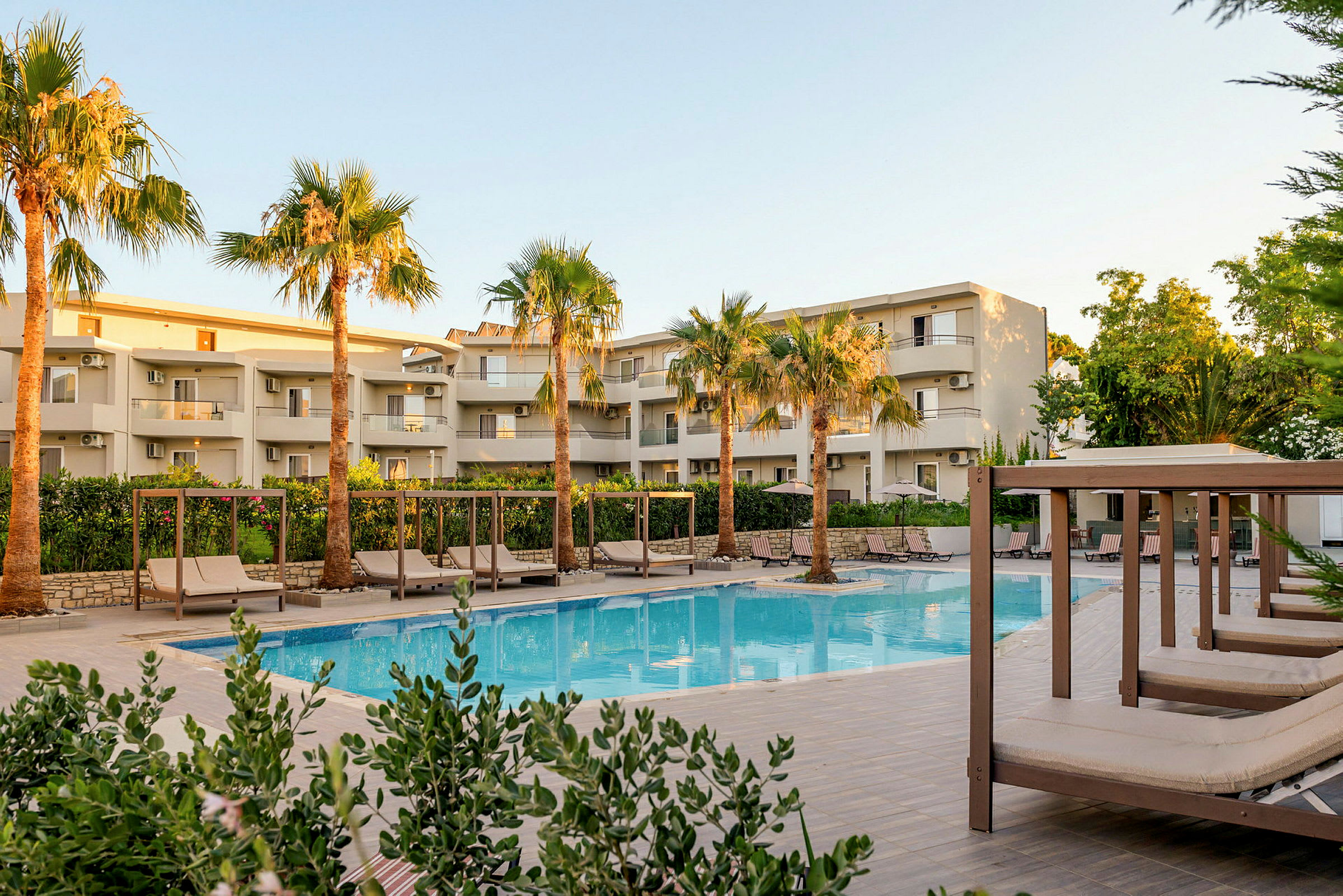 Bild av en modern hotellbyggnad med balkonger omgiven av gröna palmer. Framför byggnaden finns en pool och flera solbäddar under skuggtak. (AI-genererad text)