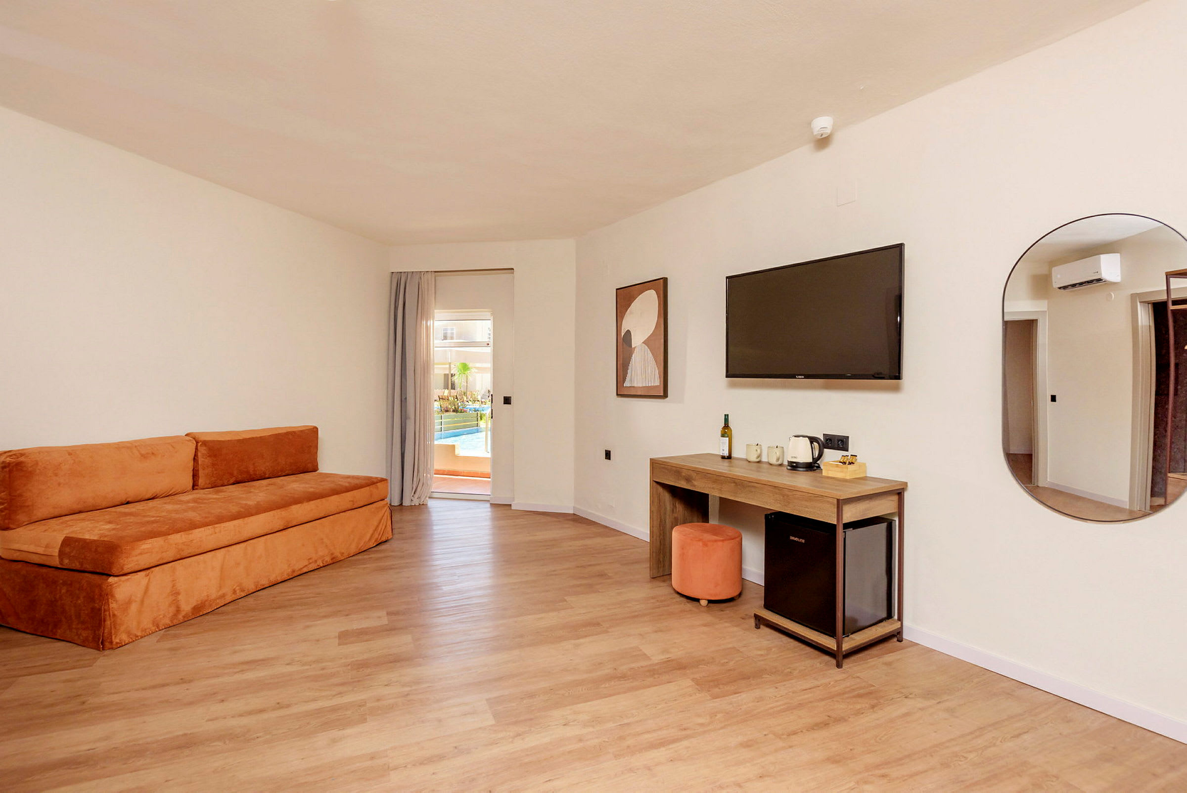 Ett hotellrum med en orange soffa, ett skrivbord, en väggmonterad TV och en dörr mot en terrass med pool. (AI-genererad text)