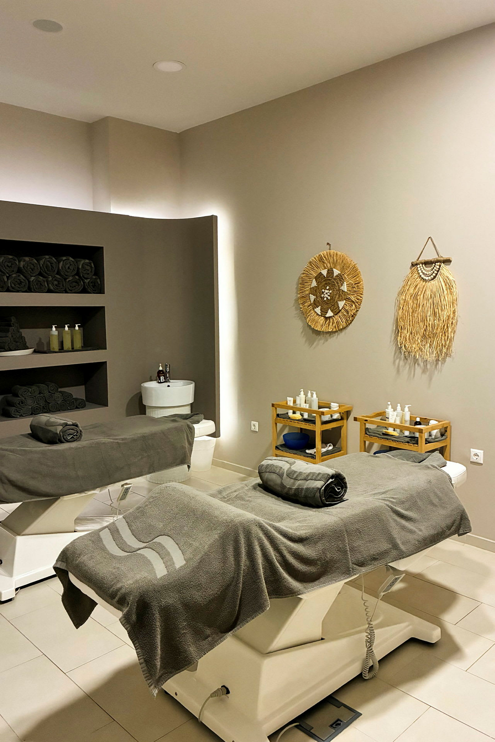 Ett spa-rum med två behandlingsbänkar klädda i grå frottéhanddukar. Väggarna är dekorerade med naturinspirerade konstverk, och hyllor med mörka handdukar och produkter syns i bakgrunden. (AI-genererad text)