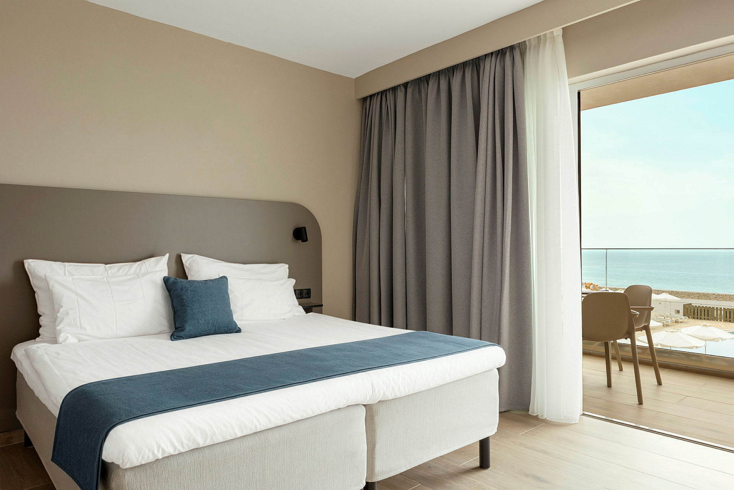 Ett hotellrum med en säng som har vita lakan och blå detaljer. Det finns en stor fönsterdörr med utsikt mot havet. (AI-genererad text)