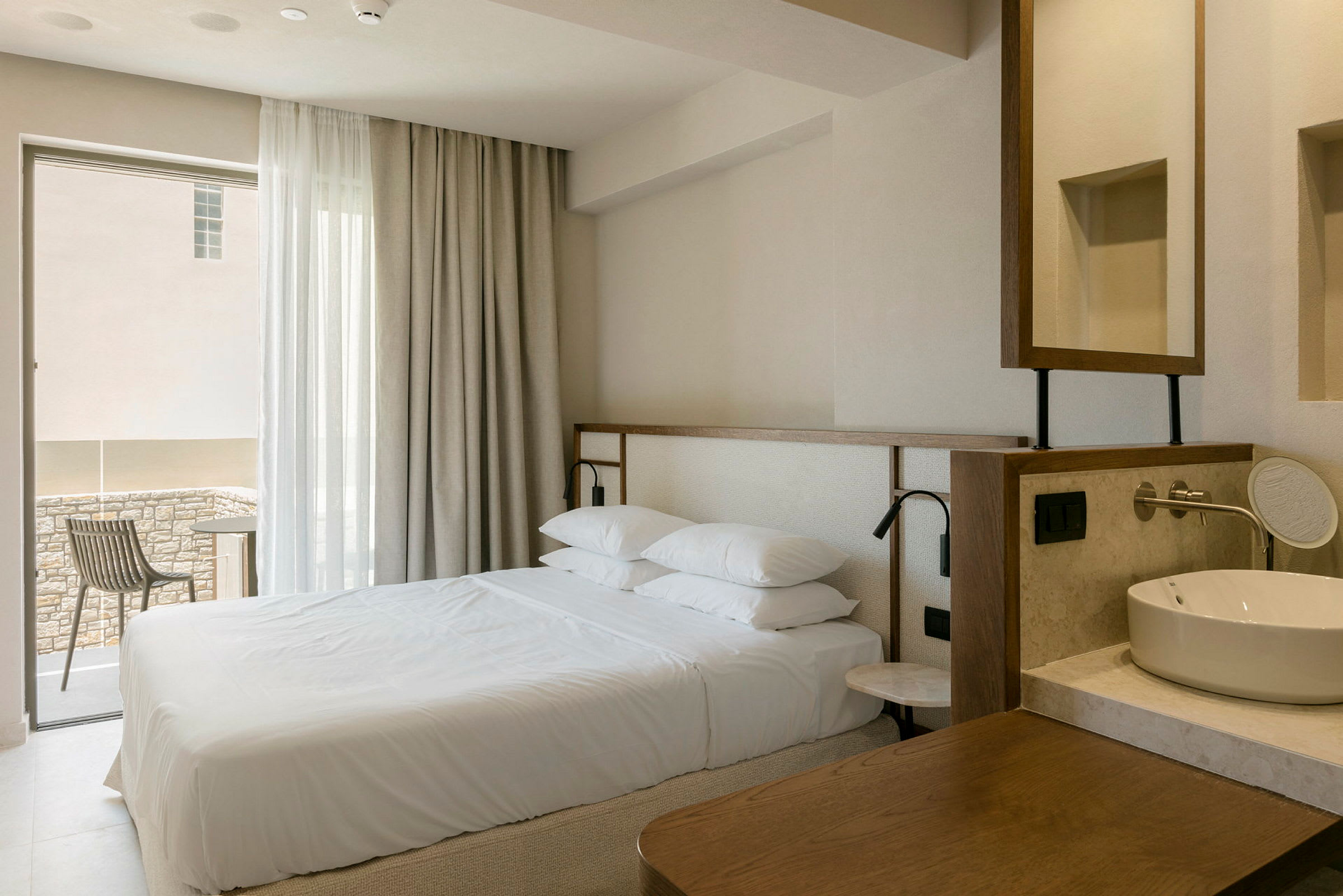 Ett hotellrumsinteriör med en dubbelsäng, ett fönster med gardiner, en balkong med stol och ett tvättställ. (AI-genererad text)