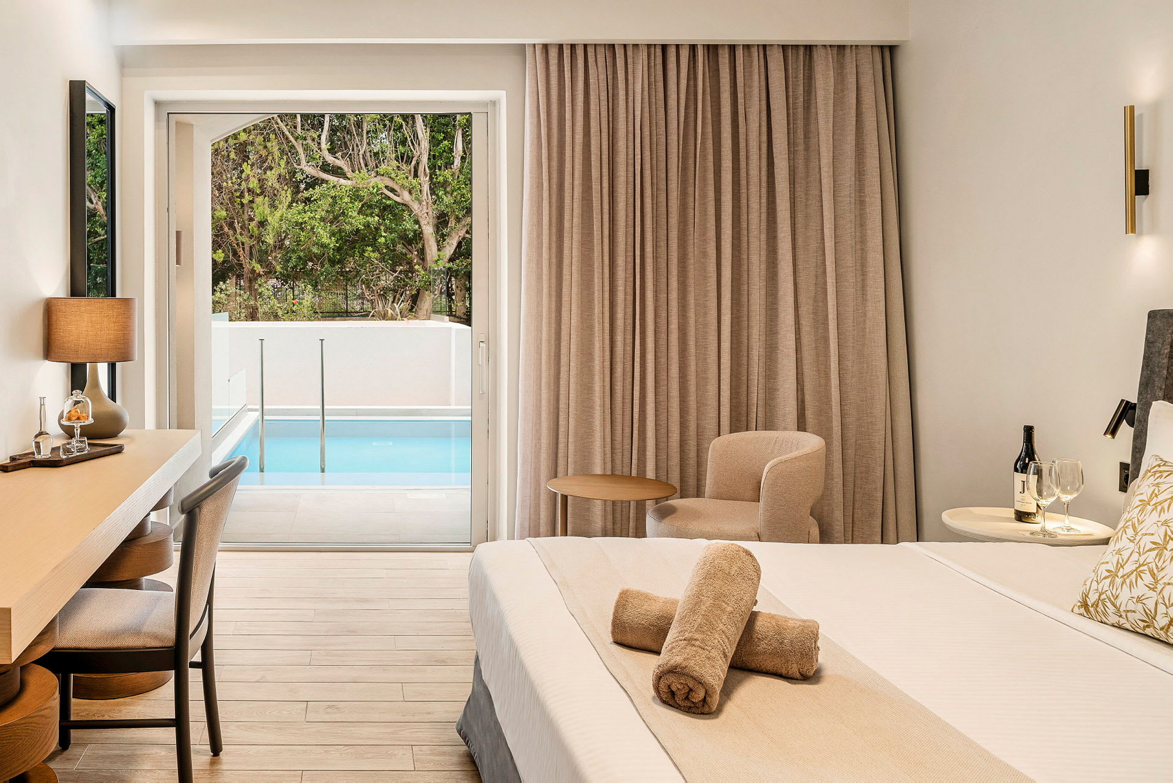 Ett hotellrum med en säng, skrivbord, samt en liten sittgrupp. Rummet har ett stort fönster som vetter mot en liten pool och grönska utomhus. (AI-genererad text)