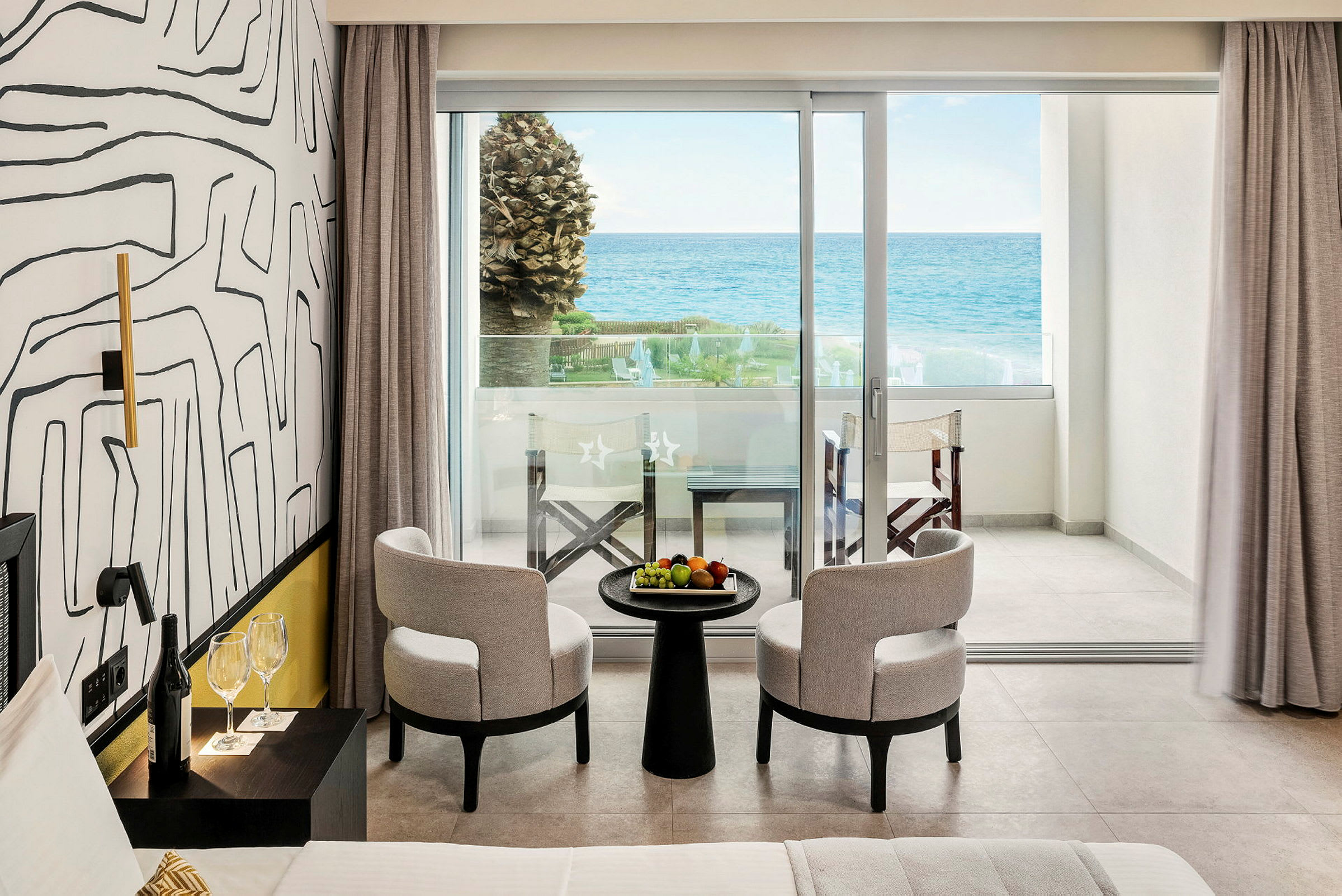 Ett hotellrum med två stolar och ett bord med frukt, samt en balkong med utsikt över havet. (AI-genererad text)