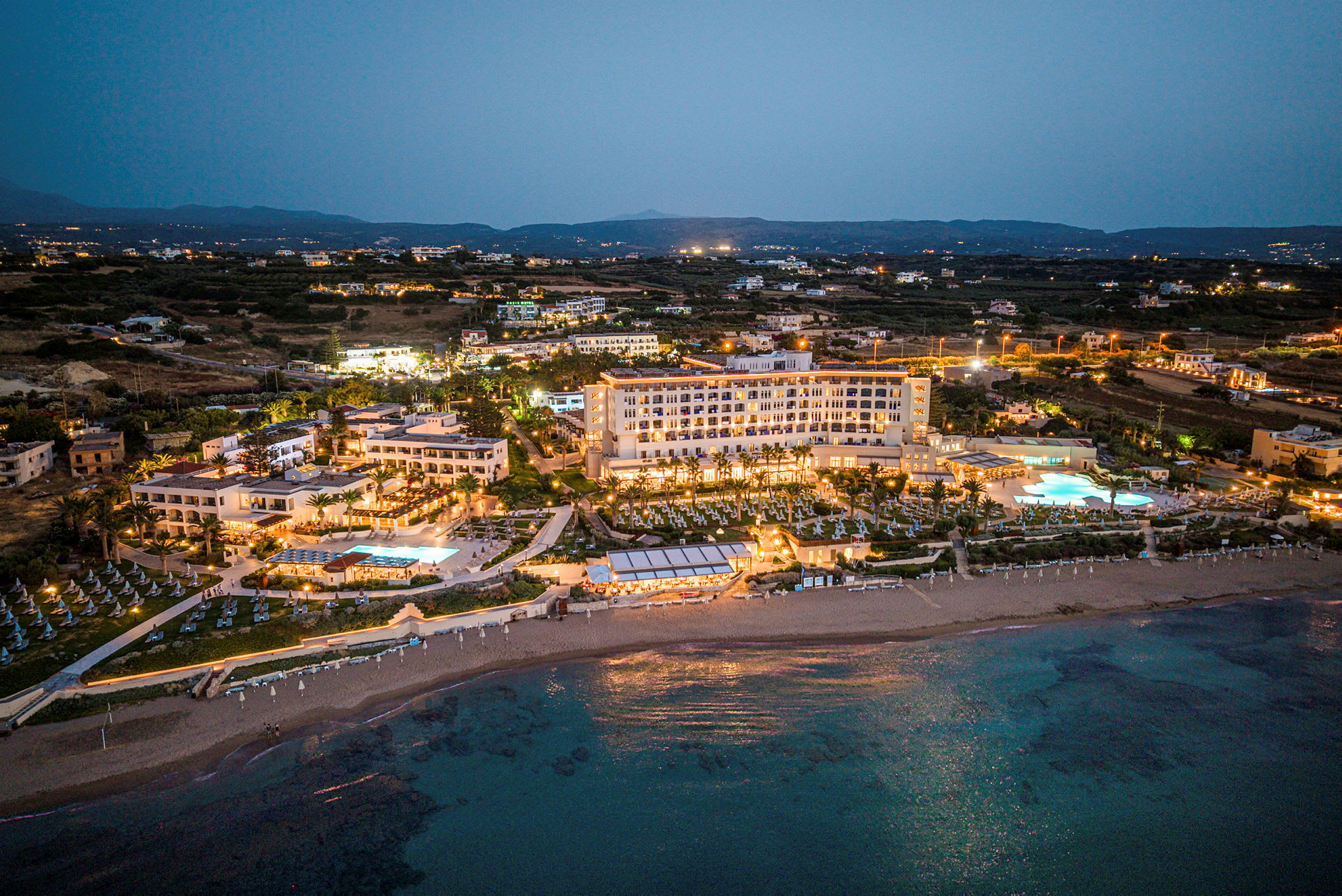 Ett hotellkomplex belyst på kvällen med pool- och strandområden synliga samt omgivande landskap i bakgrunden. (AI-genererad text)
