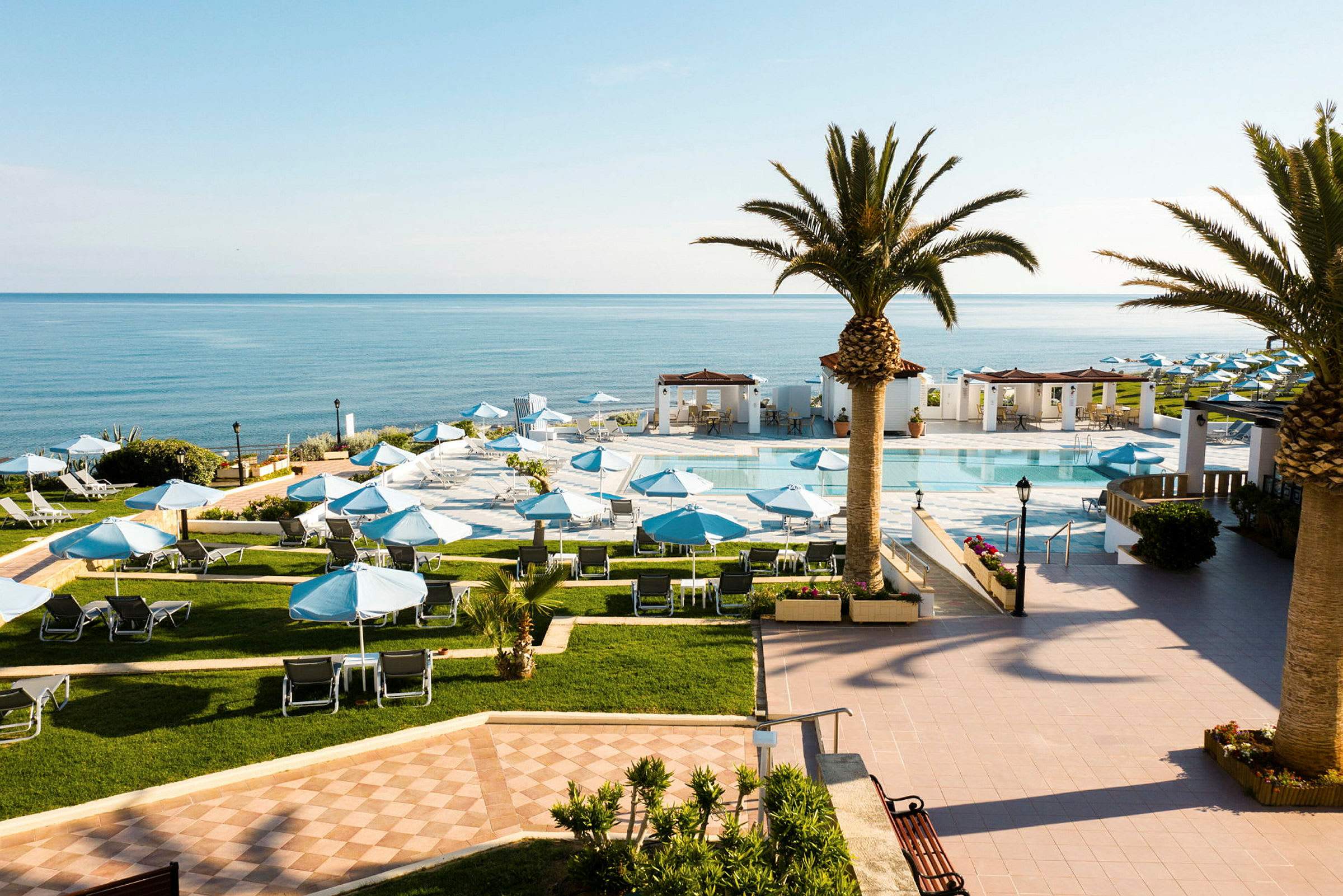 Ett hotellområde med en pool och solstolar under blå parasoller, omgärdad av palmer och med utsikt över havet. (AI-genererad text)