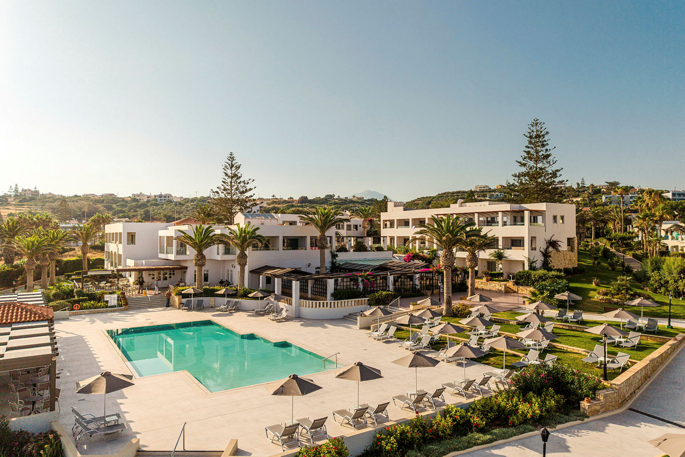 En översiktsbild av ett semesterresort med en pool, solstolar med parasoller samt omgivande byggnader och grönska. (AI-genererad text)