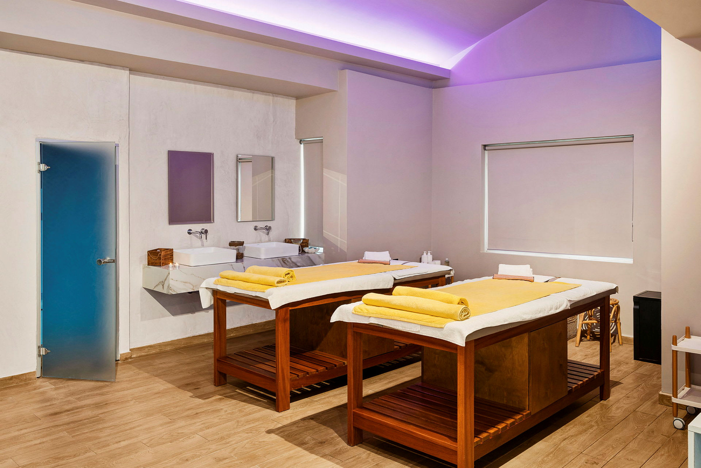 Ett spa-rum med två massagebänkar utrustade med gula handdukar. Det finns två handfat med speglar på väggen och en blå dörr på sidan. (AI-genererad text)
