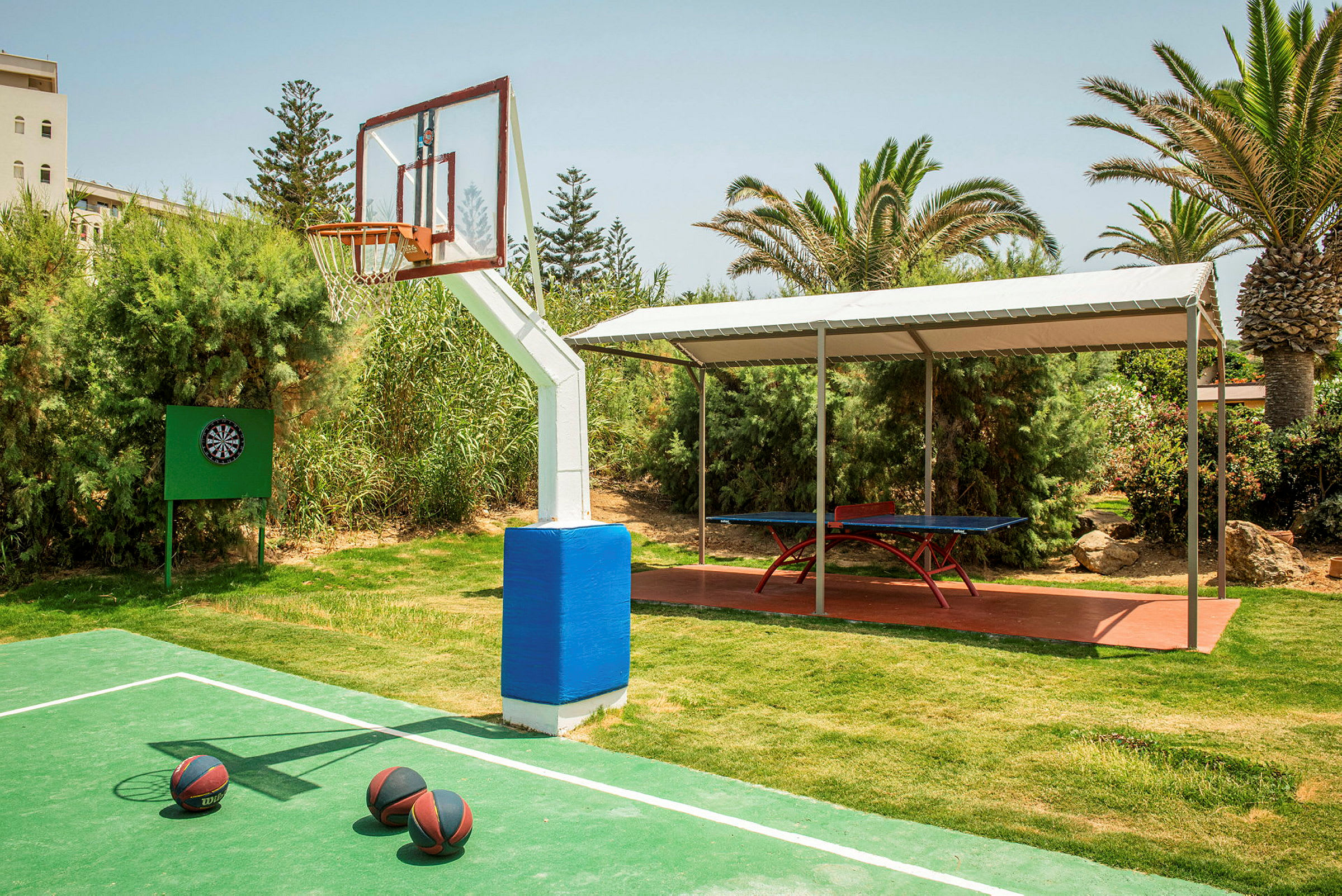 En utomhusbasketplan med tre basketbollar på marken framför en basketkorg. Ett darttavla är också i sikte på vänster sida, och i bakgrunden finns ett bordtennisbord under ett tak. (AI-genererad text)