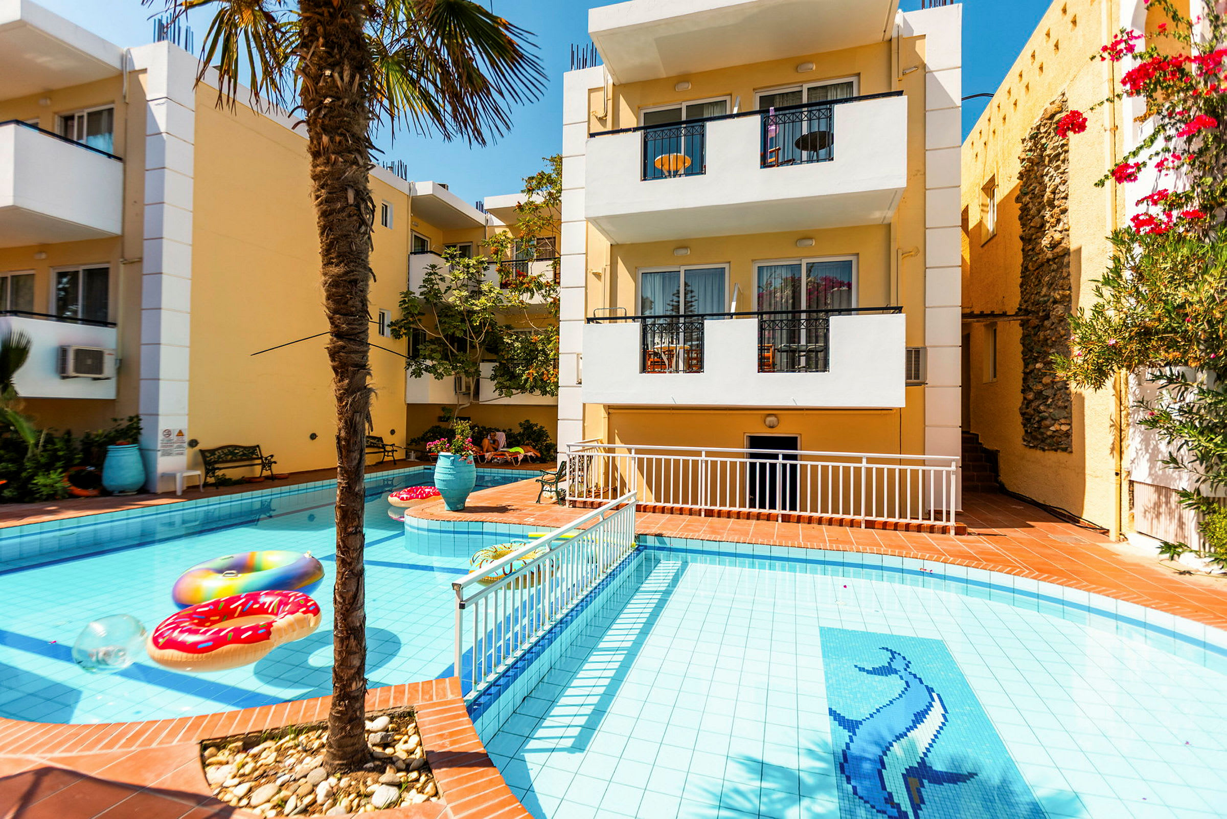 Bild av en byggnad med en pool framför. Poolen har en dekoration av en delfin och flytande föremål, samt omges av en terrass med växter och utemöbler. (AI-genererad text)