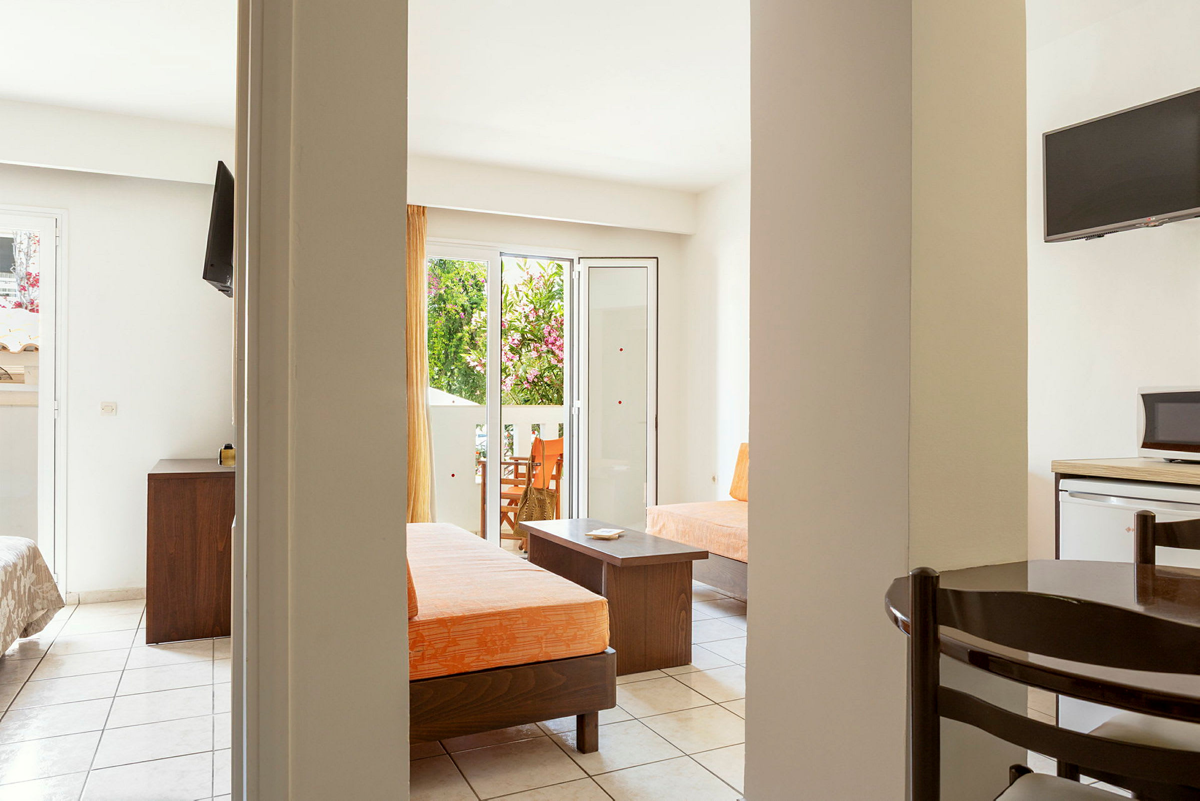 Ett hotellrum med öppen planlösning, möblerat med orange dynor, bord och stolar samt en TV. Dörren till en balkong som vetter mot grönska syns i bakgrunden. (AI-genererad text)