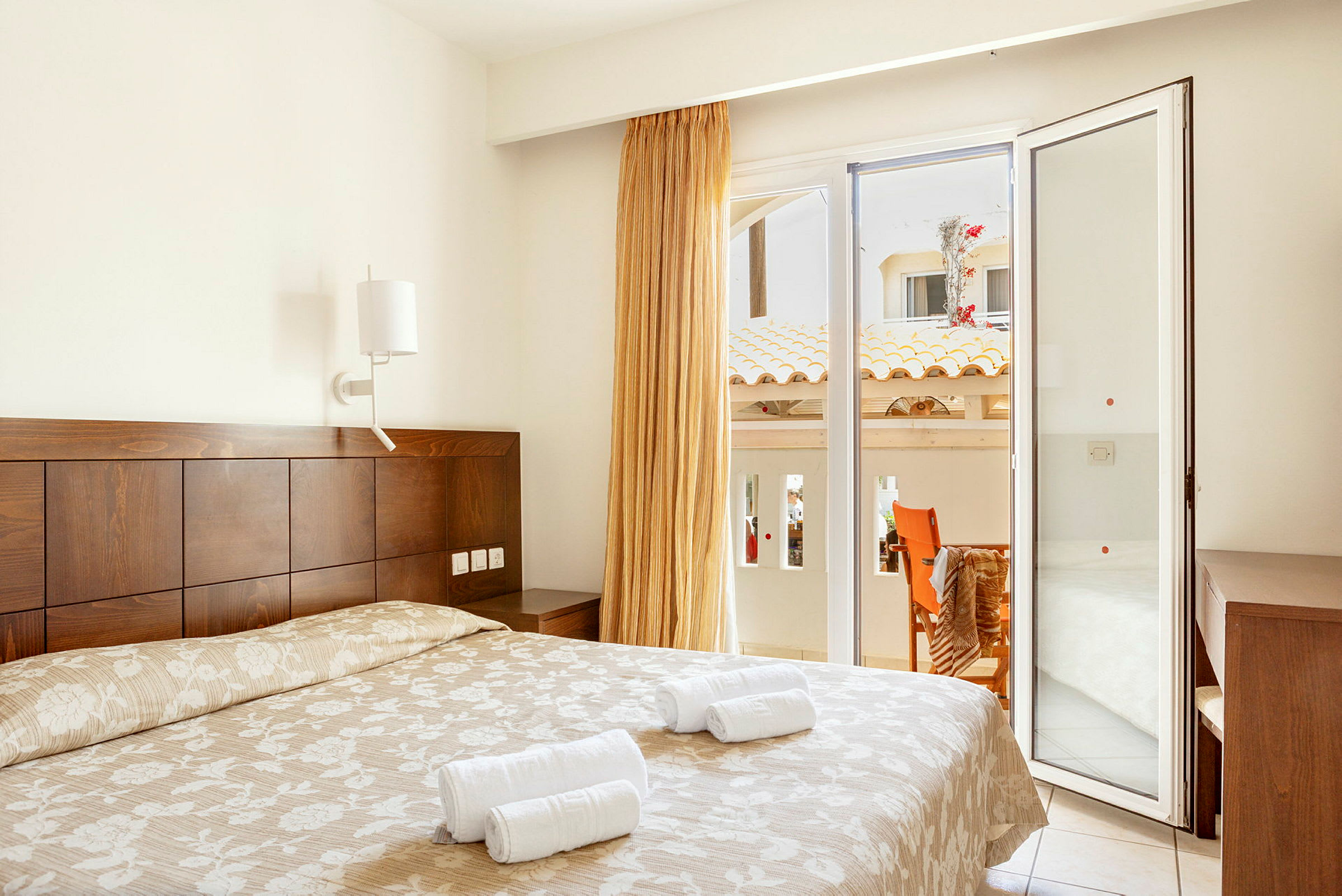 Ett hotellrum med en dubbelsäng, ljust täcke och handdukar ovanpå. Vid sidan finns en glasdörr som leder ut till en balkong med utsikt över byggnader. (AI-genererad text)