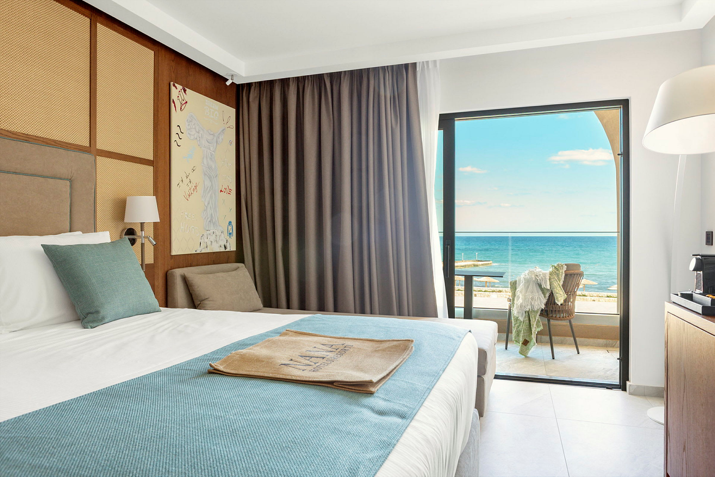 Hotellrum med en bäddad säng klädd i vit och blå sängklädsel. Rummet har stora fönster med utsikt över havet och en möblerad balkong. (AI-genererad text)