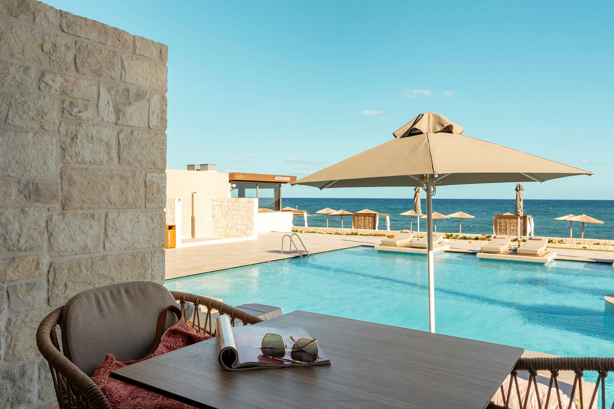 Utsikt från en altan mot en pool med parasoller och solstolar, med havet i bakgrunden. (AI-genererad text)