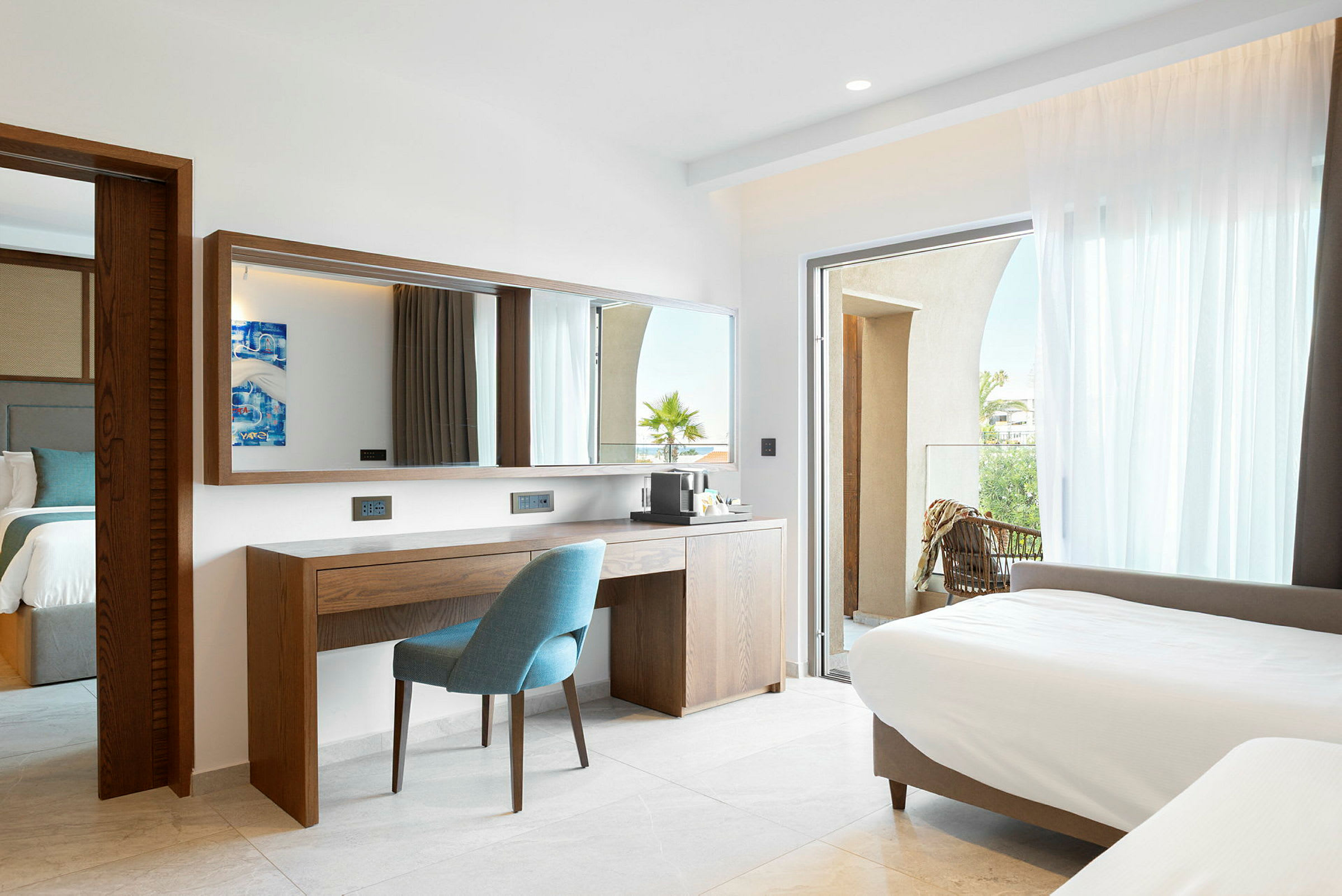 Ett hotellrum med en modern inredning, inklusive en säng, ett skrivbord med en stol och en spegel. Rummet har en balkong med utsikt. (AI-genererad text)