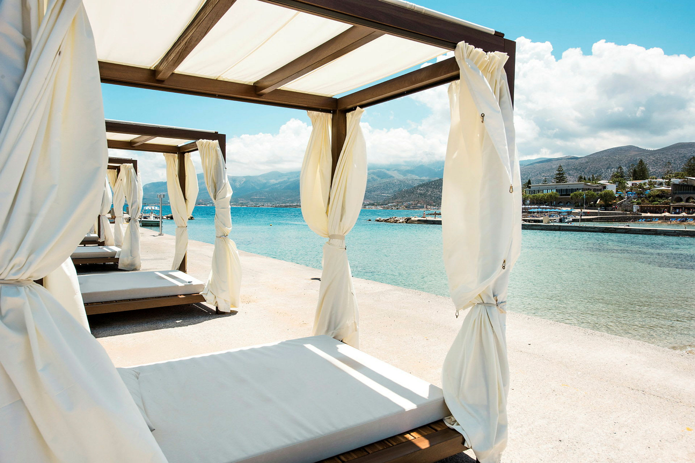 Solbäddar med vita draperier vid strandkanten med hav och berg i bakgrunden. (AI-genererad text)