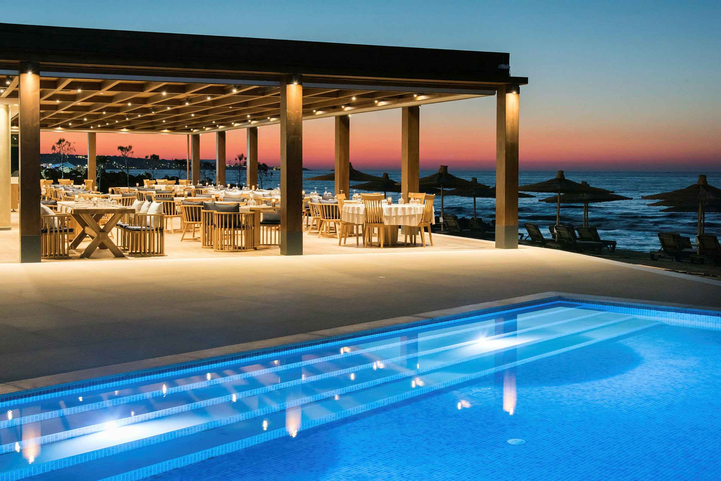 Restaurang med bord och stolar nära en pool och havet vid solnedgång. (AI-genererad text)